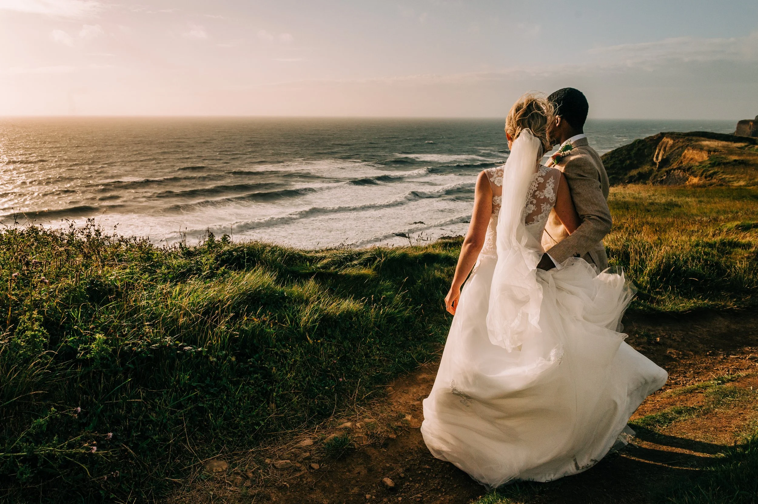 A bride and groom sharing a kiss on a grassy cliff overlooking the ocean at sunset.