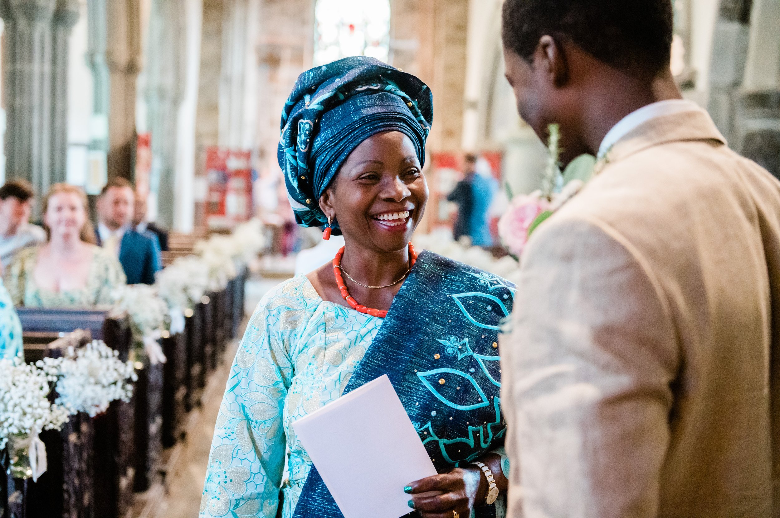 A smiling woman wearing a blue headwrap and traditional African attire, holding a white document, standing inside a church during a wedding ceremony, with guests seated behind her.