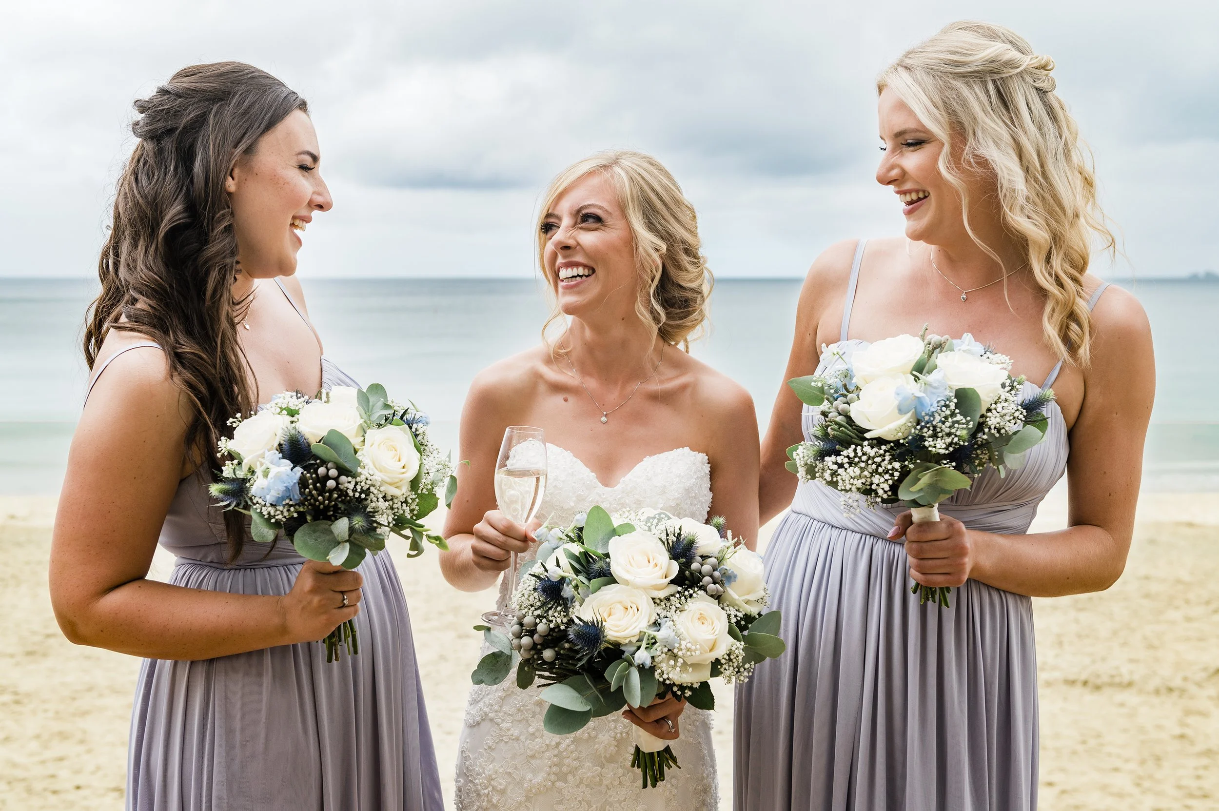 A bride and two bridesmaids smiling and holding bouquets on the beach.