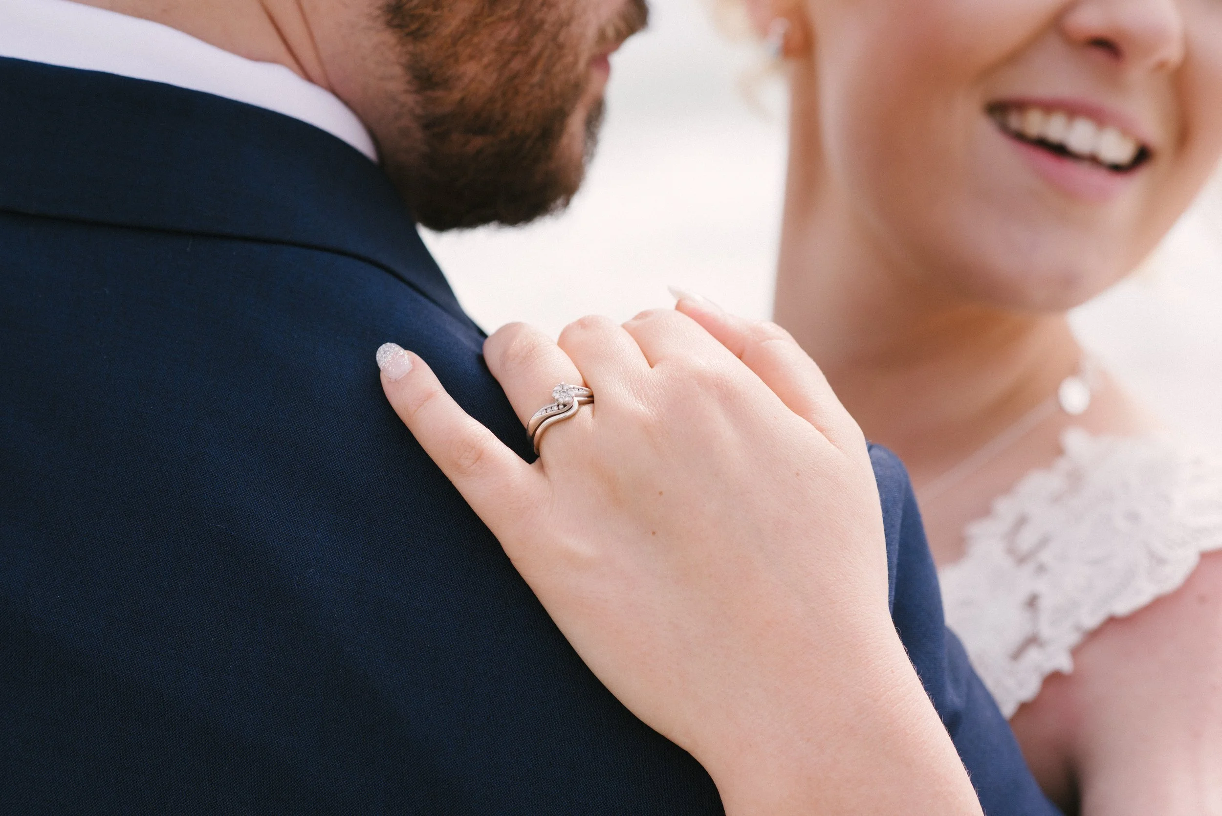 Close-up of a bride's hand resting on a groom's shoulder, showing a wedding ring, with part of the bride's face smiling in the background.