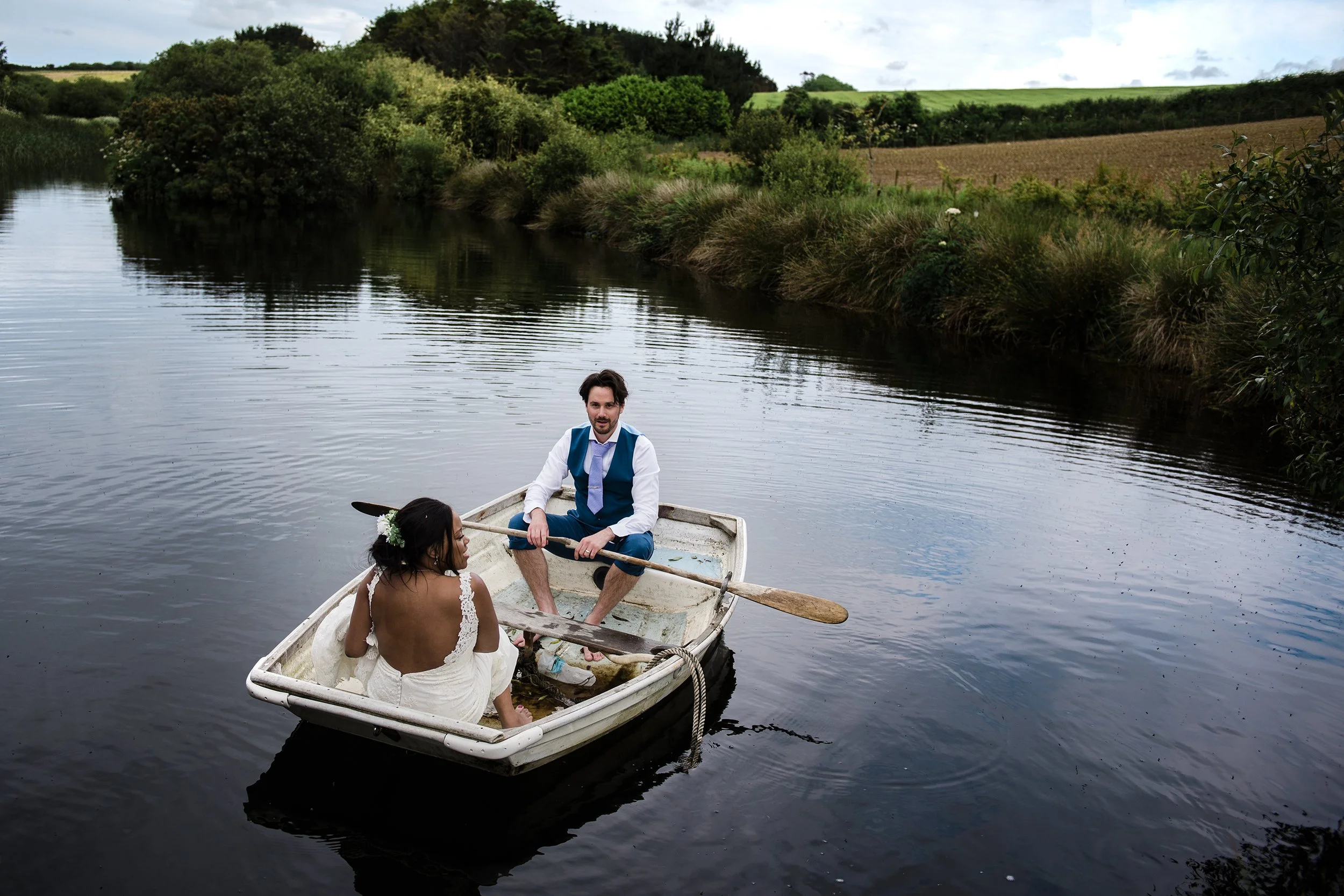A couple in wedding attire sitting in a small rowboat on a calm river, surrounded by lush green trees and fields, with a cloudy sky overhead.