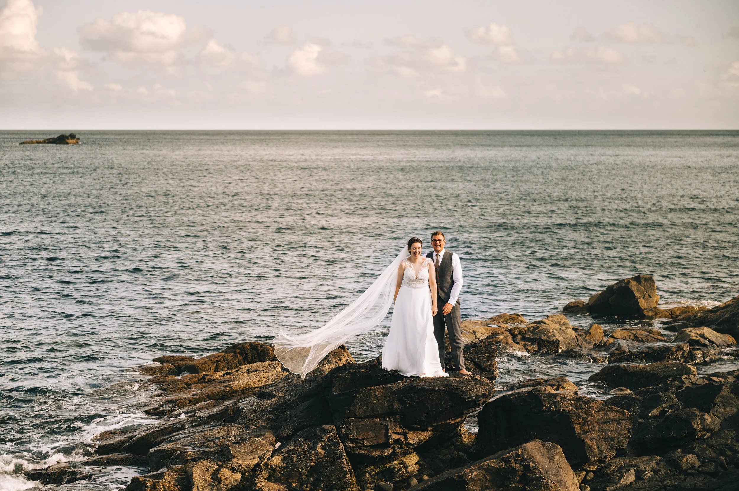 A bride in a white wedding dress and a groom in a gray vest and pants celebrating on rocks by the ocean during sunset.