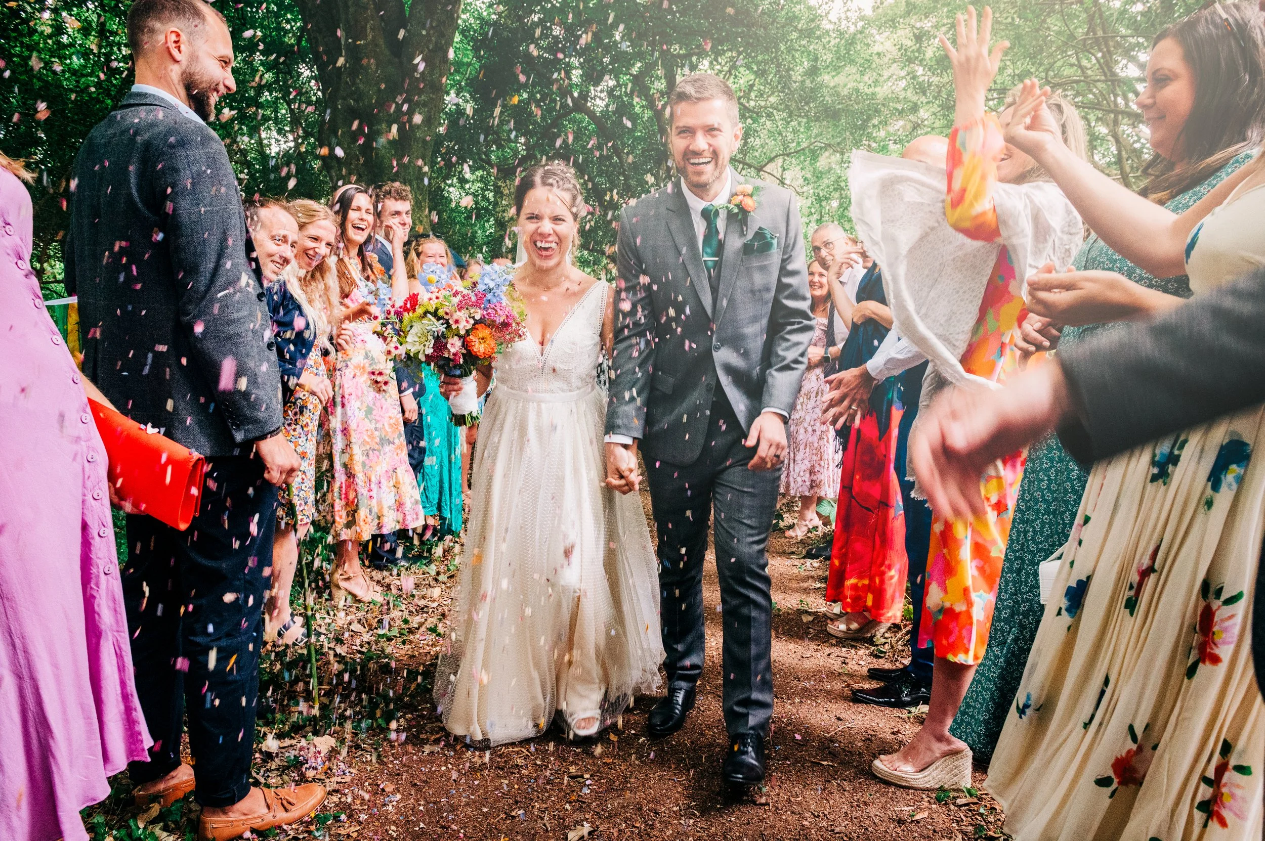 A happy newlywed couple walking hand-in-hand through a crowd of celebrating guests, who are throwing confetti and cheering. The bride wears a white dress and holds a colorful bouquet, and the groom is in a gray suit with a tie.