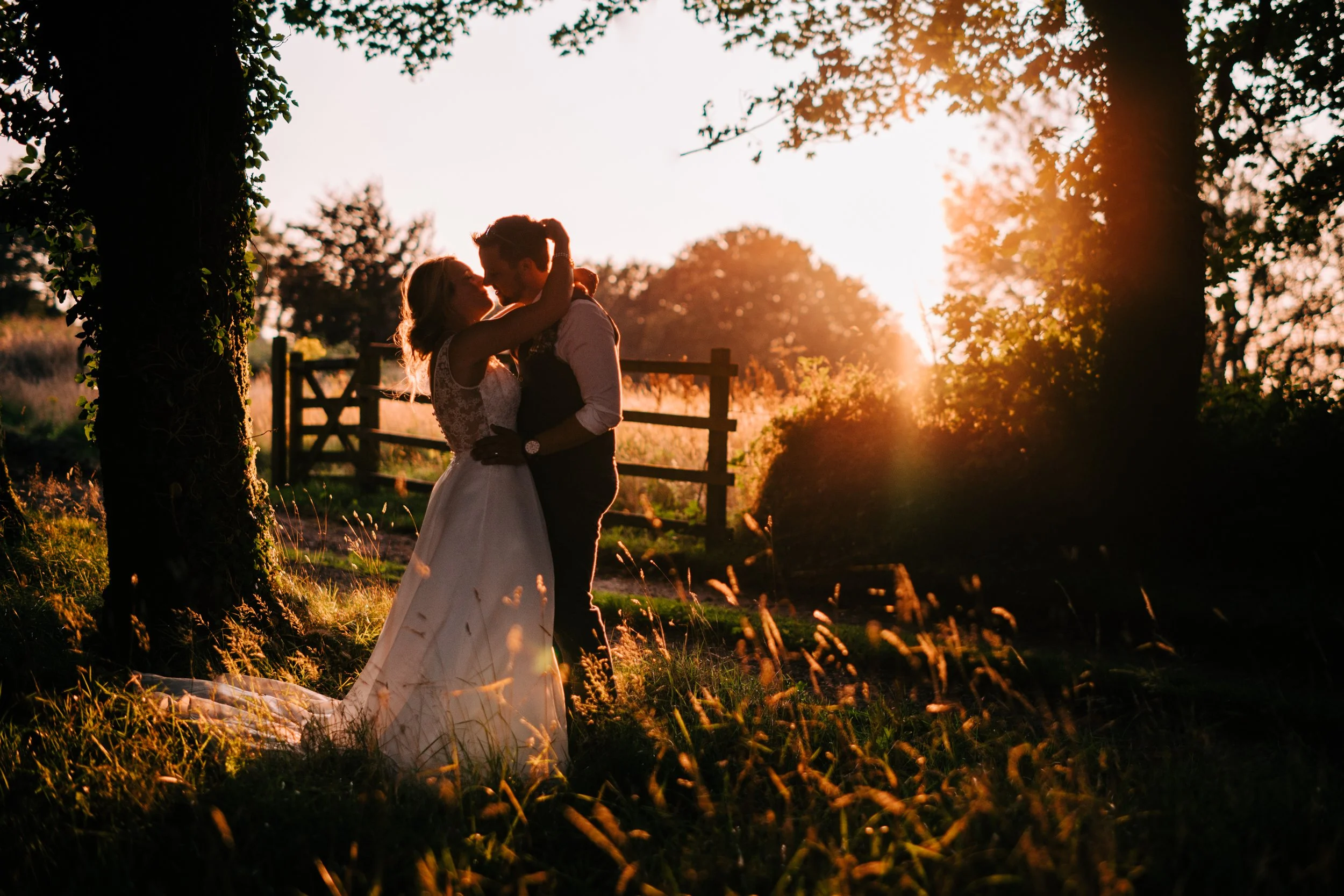 A silhouetted couple kisses outdoors during sunset, surrounded by trees and grass, with a wooden fence in the background.