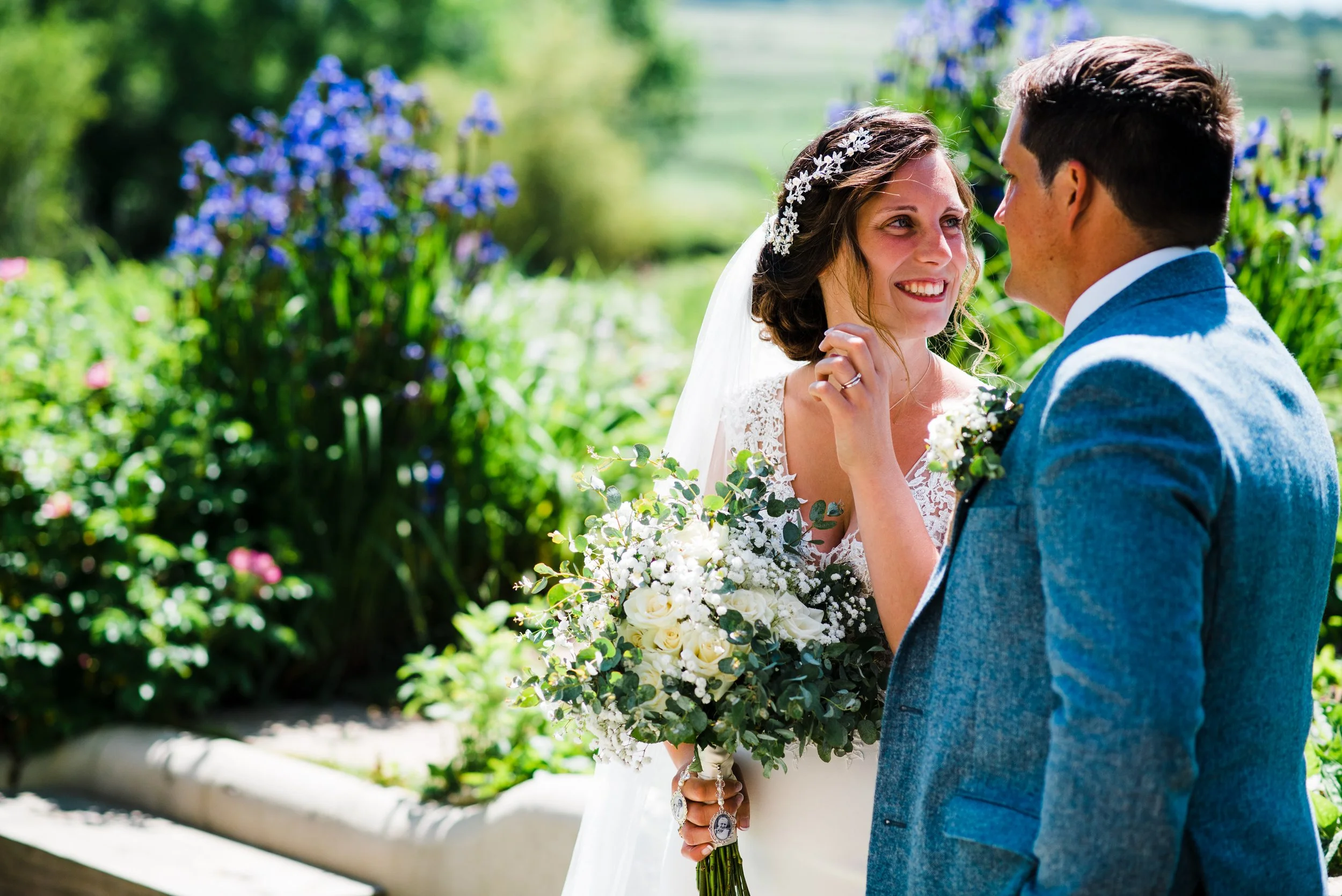 A bride and groom at their wedding outdoors, looking at each other. The bride holds a bouquet of white flowers and wears a lace dress with an intricate headpiece. The groom wears a blue suit. The background features greenery and blooming flowers.
