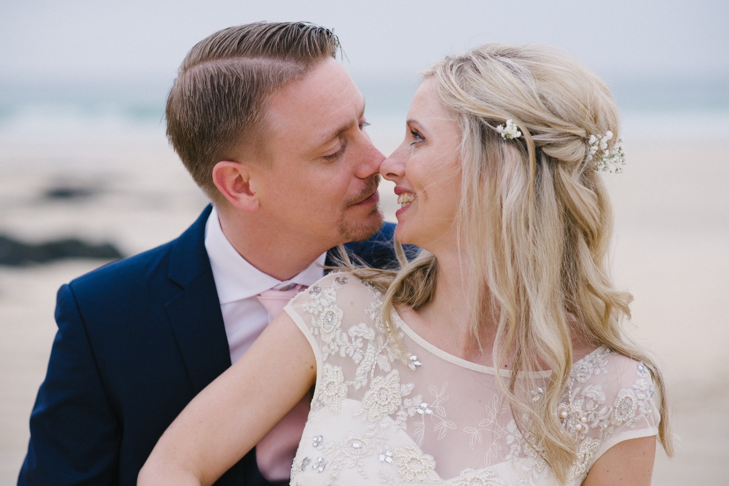 A bride and groom close together on a beach, with faces near touching, smiling, and looking into each other's eyes.
