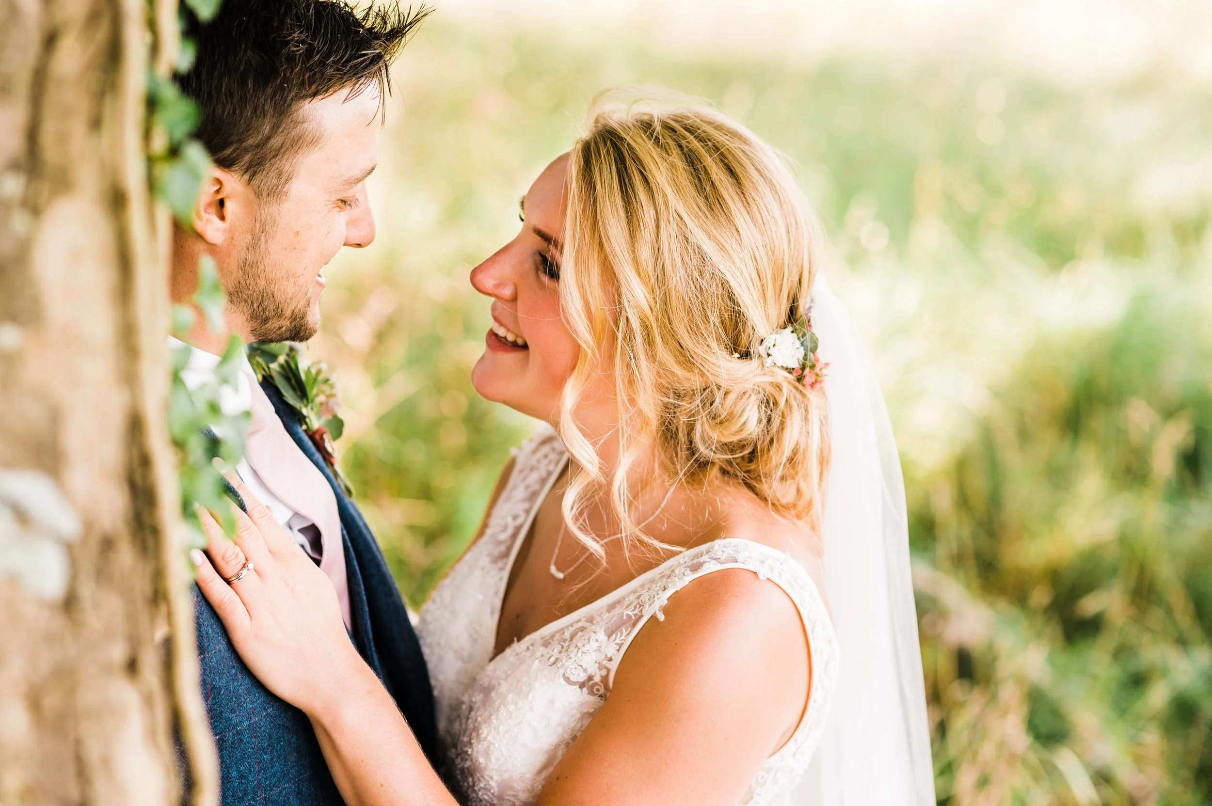 A bride and groom smiling at each other outdoors near a tree in a wedding setting.