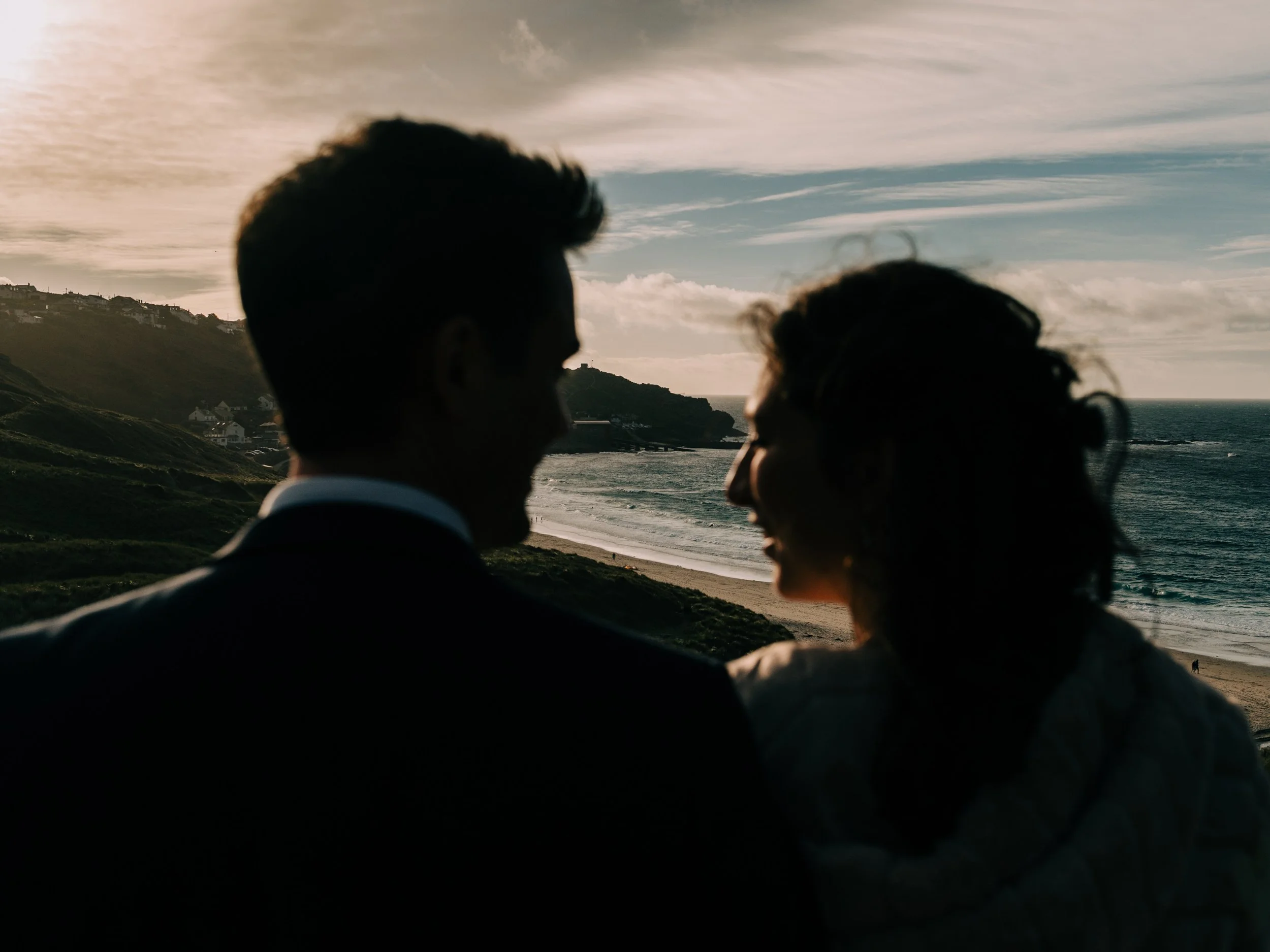Silhouette of a couple facing each other on a beach during sunset, with waves and hills in the background.