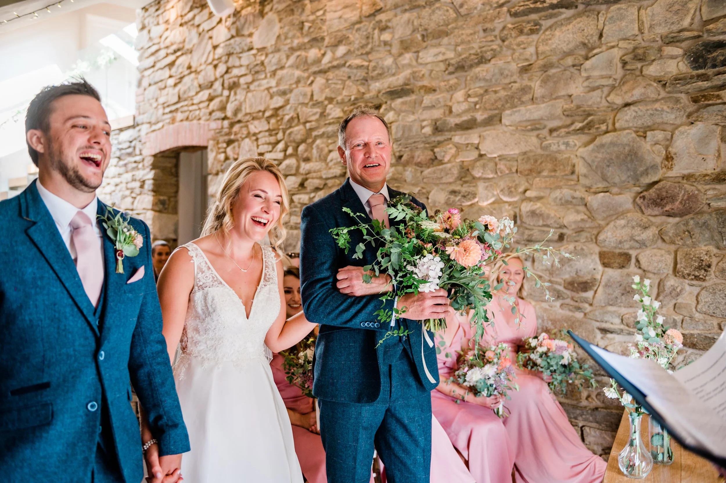 Wedding ceremony with bride, groom, and wedding party. Bride and groom holding hands, bride smiling, groom holding bouquet of flowers. Bridesmaids in pink dresses sitting with bouquets. Stone wall background.
