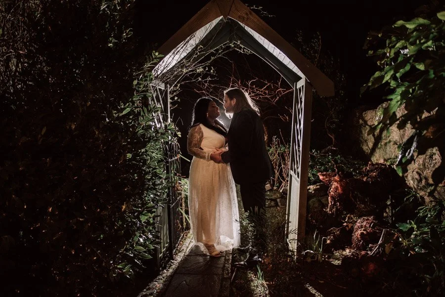 A couple standing under a small white garden archway at night, holding hands and facing each other.