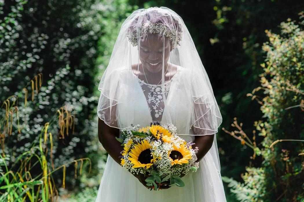A bride in a white wedding dress with a sheer veil holding a bouquet of sunflowers and white flowers, standing outdoors in a lush green garden.