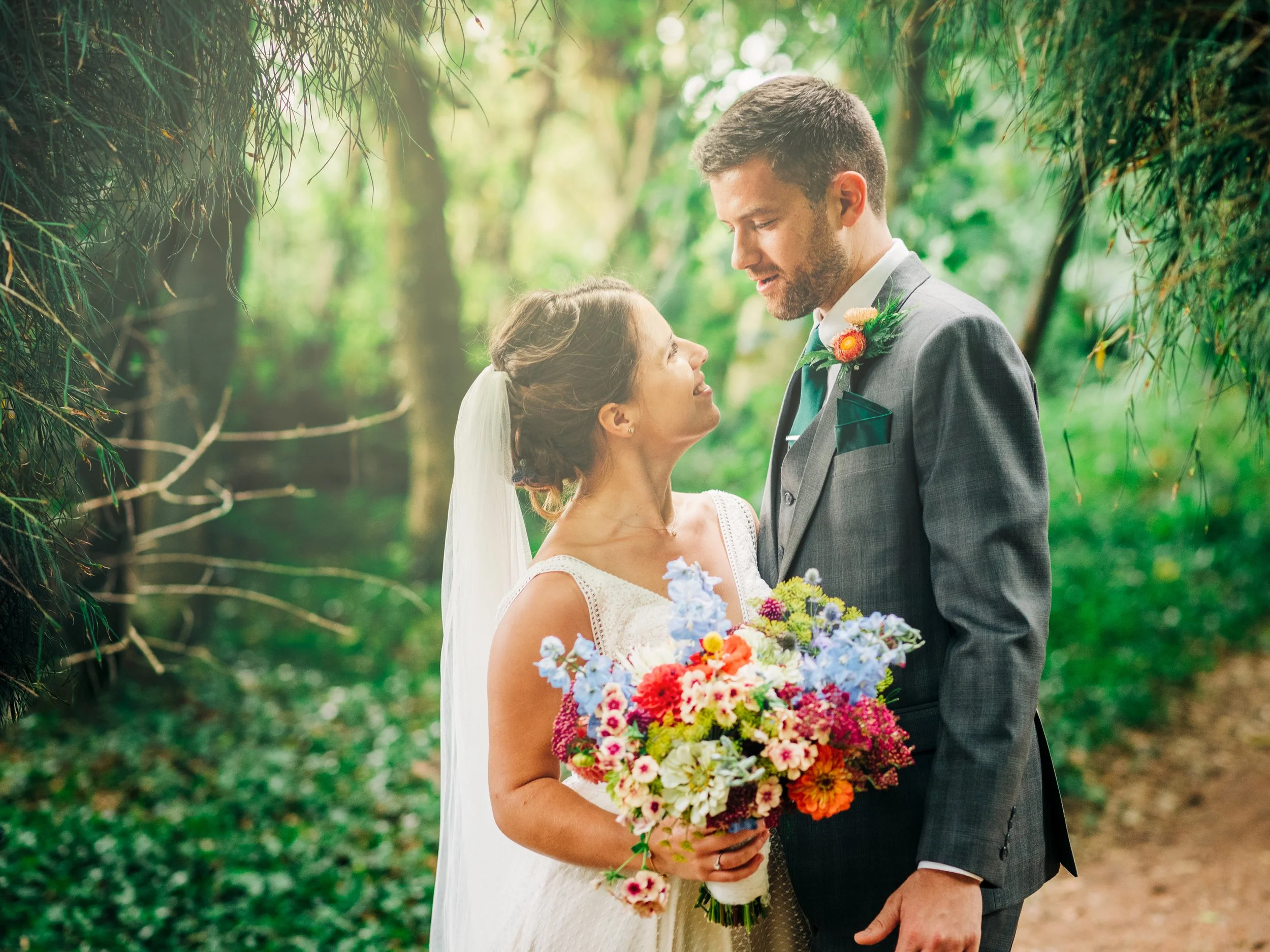 A bride and groom stand close together in a lush, green outdoor setting, gazing into each other's eyes. The bride holds a colorful bouquet of flowers, and the groom wears a gray suit with a boutonniere. Soft sunlight filters through the trees.