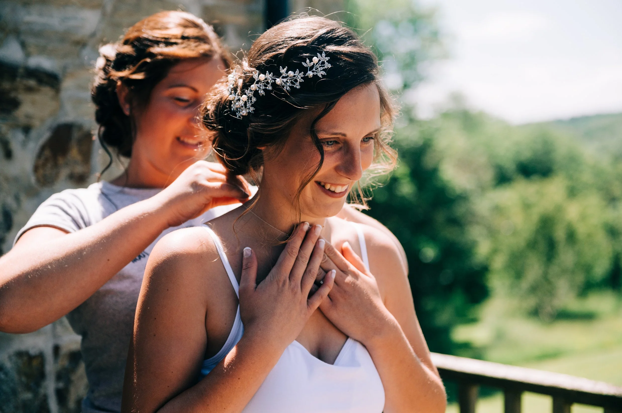 A woman in a white dress and a floral headpiece smiles as she receives help adjusting her hair from another woman in a gray t-shirt on a sunny outdoor setting with greenery in the background.