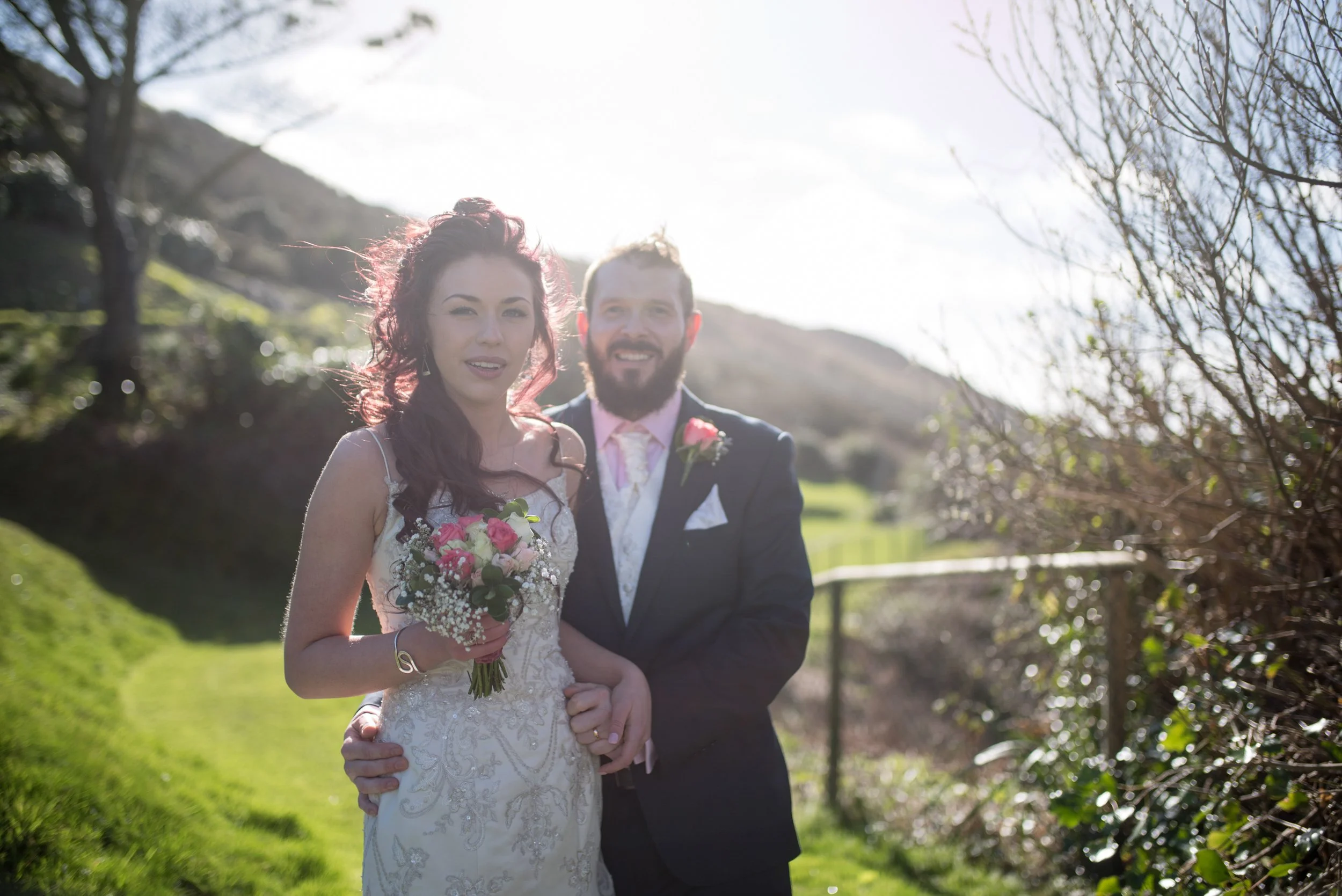 A bride and groom outdoors on a sunny day with trees and a hillside in the background, smiling and holding a bouquet of flowers.