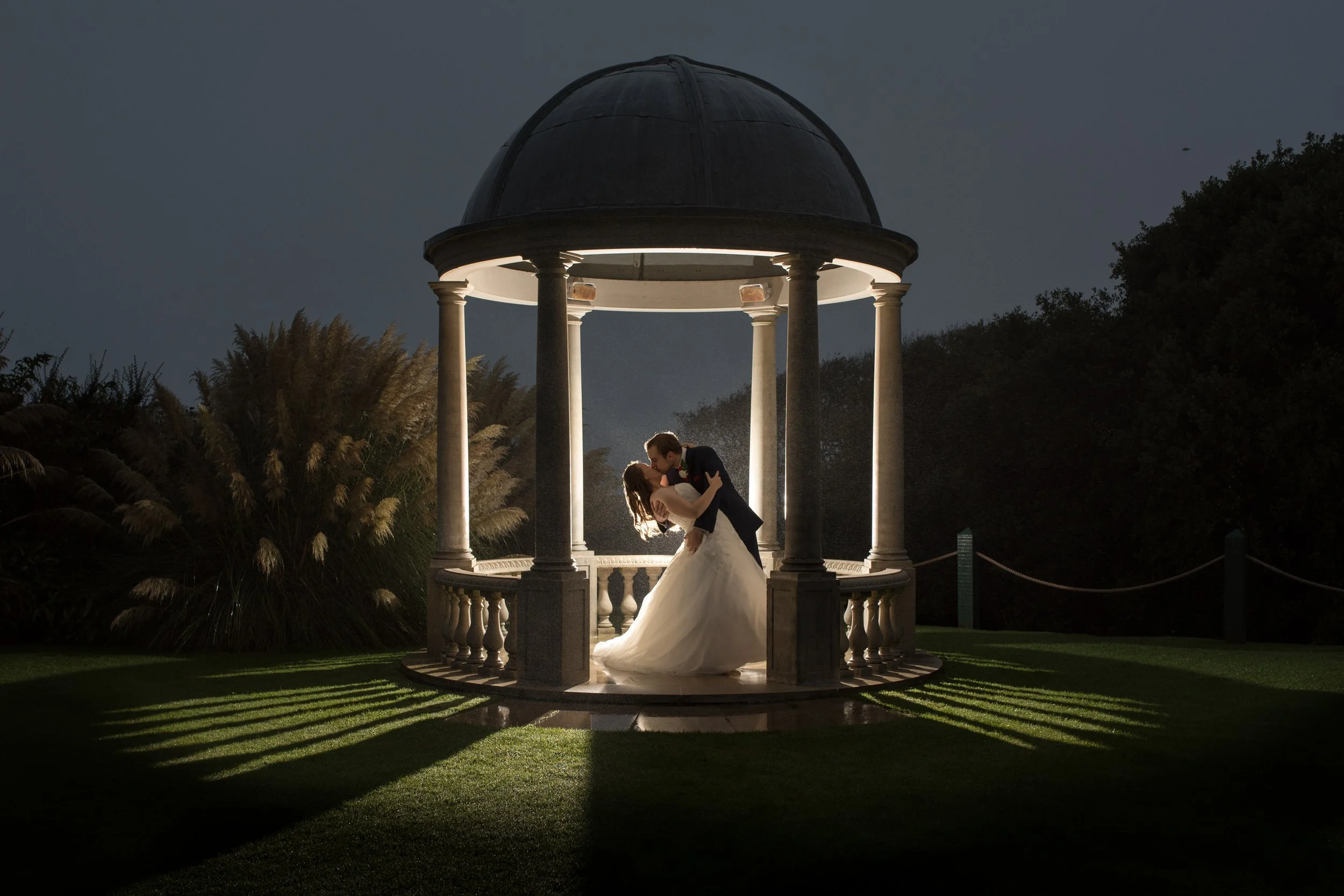 A bride and groom kissing in a circular, open-air gazebo at night, surrounded by greenery, with dramatic lighting highlighting them and casting shadows on the grass.