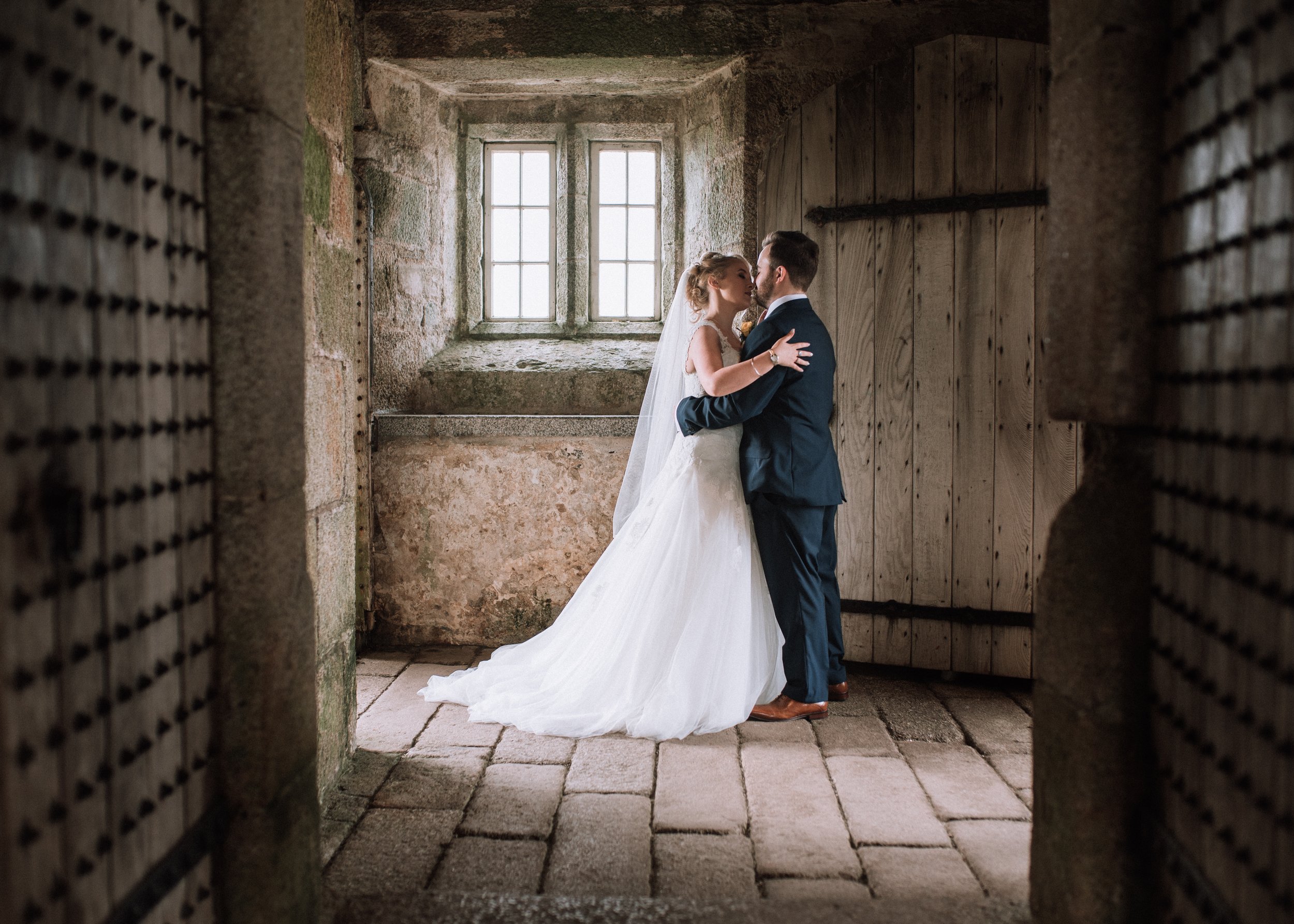A bride and groom embrace in a rustic stone and wood room with a window in the background.