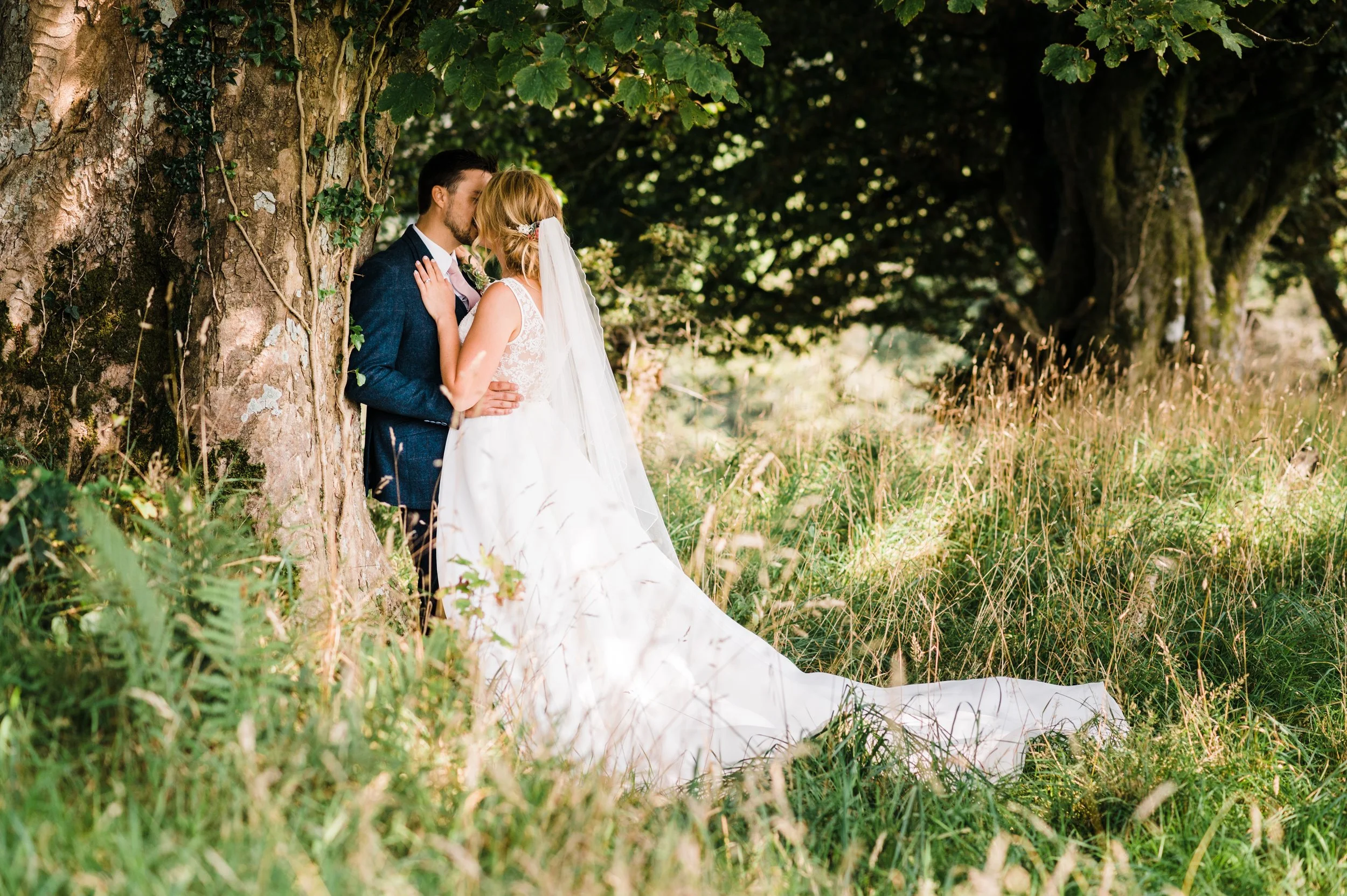 A bride and groom share a kiss outdoors next to a large tree, surrounded by tall grass and greenery, with the bride wearing a white wedding gown and veil and the groom in a dark suit.