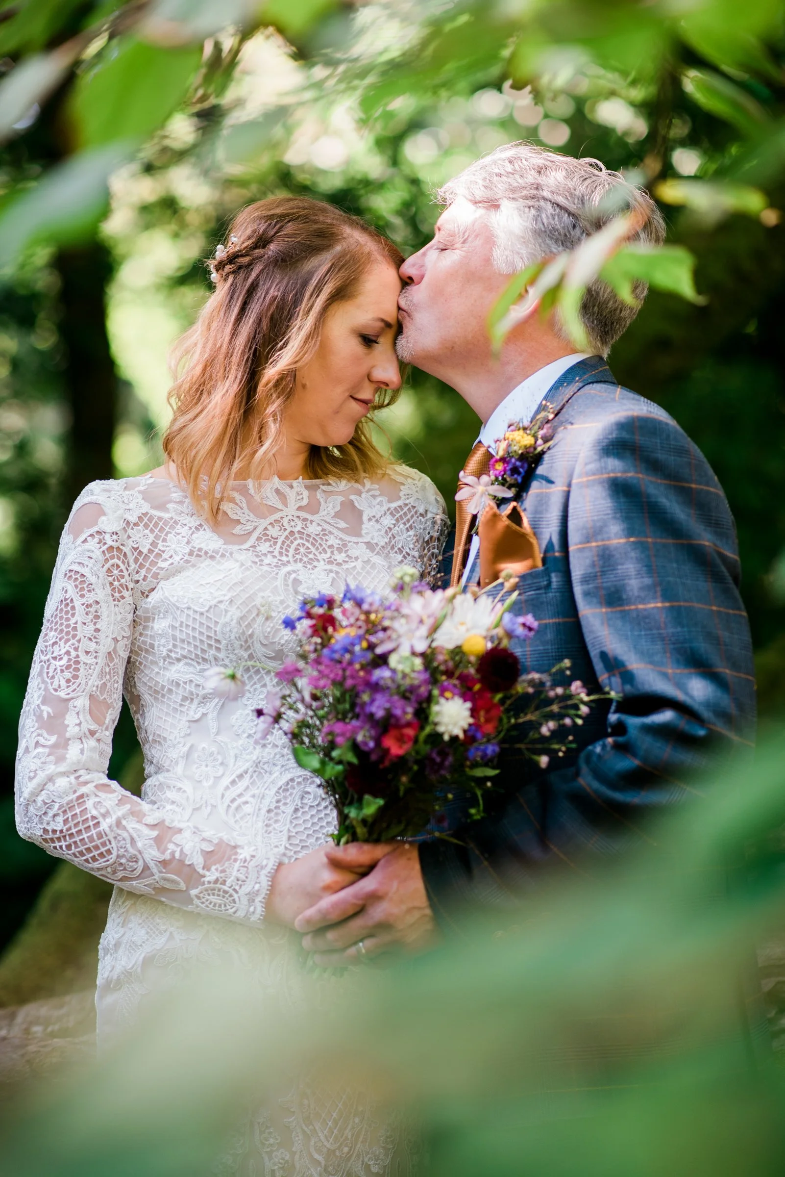 A bride and groom share a tender moment outdoors, with the groom kissing the bride on the forehead. The bride is holding a colorful bouquet of flowers, and both are dressed in wedding attire surrounded by lush green foliage.
