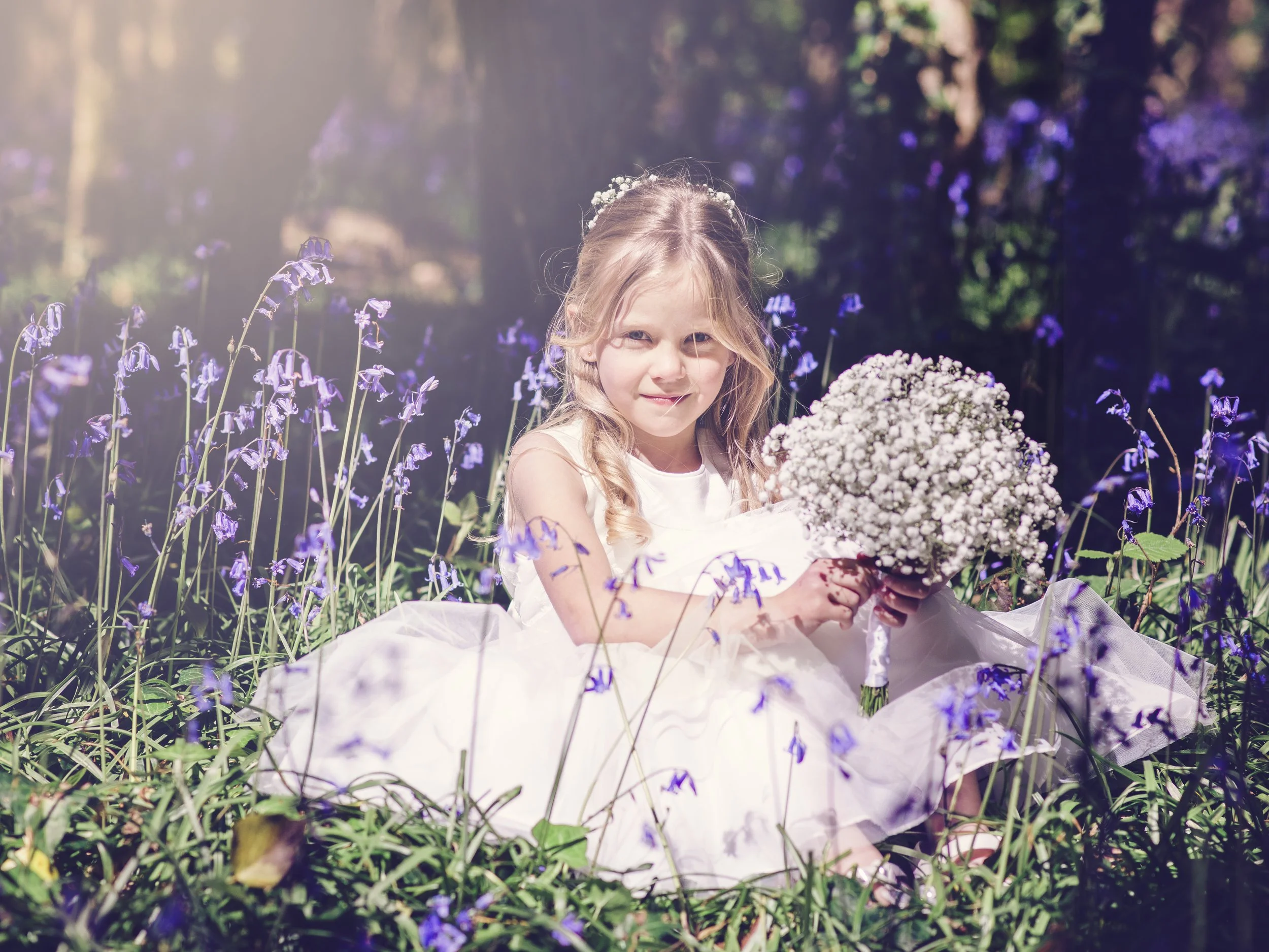 A young girl in a white dress sitting in a field of purple flowers holding a bouquet of white flowers.