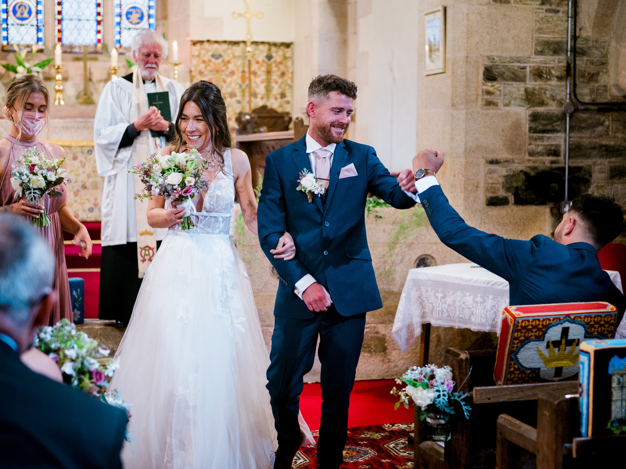 A wedding ceremony inside a church with a bride and groom smiling. The bride is holding a bouquet, the groom is showing his watch. Bridesmaids and an officiant are visible behind them.