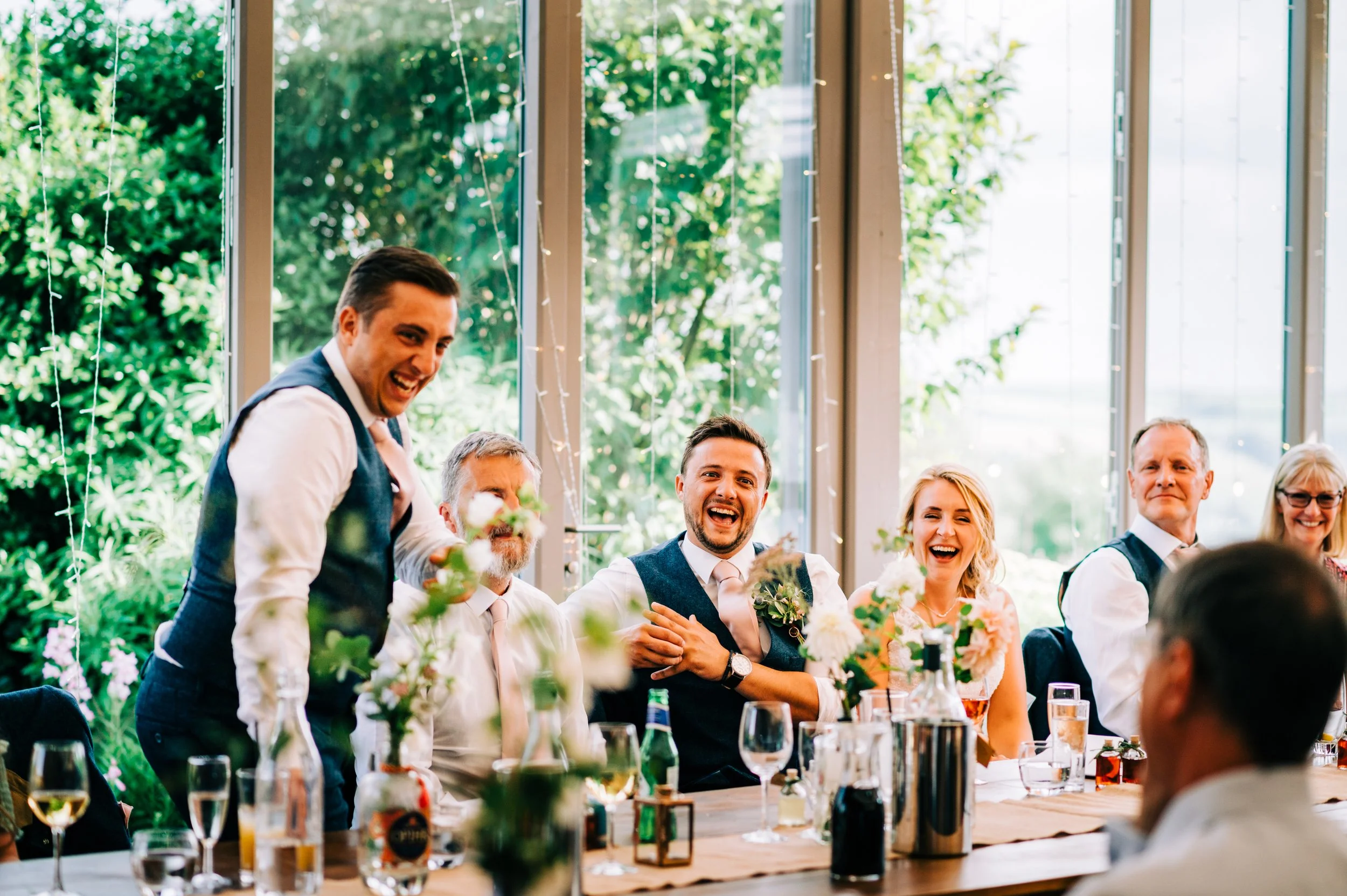 A man giving a speech at a wedding reception, with guests seated at a decorated table, smiling and laughing.