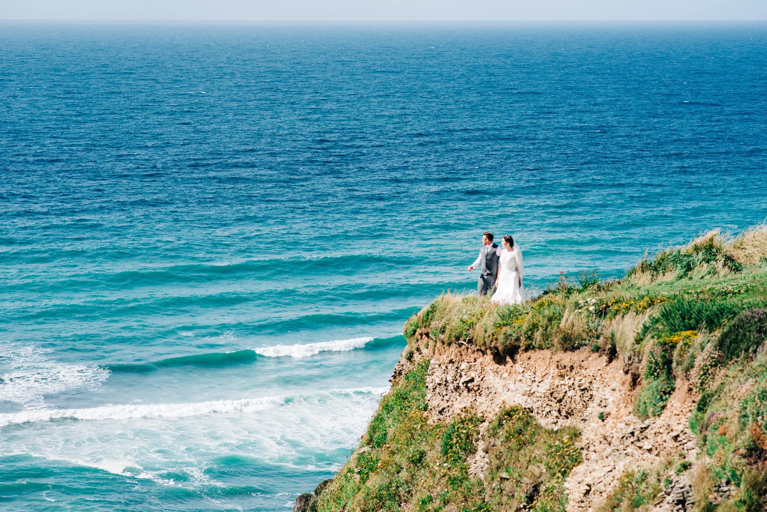 A bride and groom walking on a grassy cliffside overlooking the ocean, dressed in wedding attire.