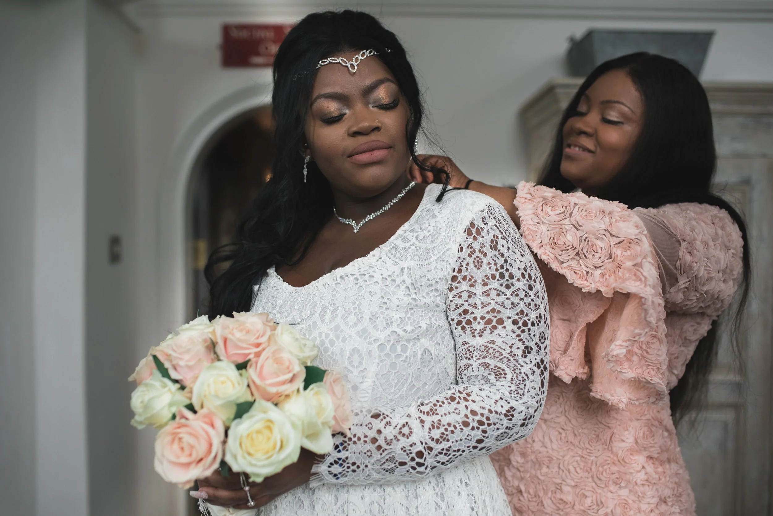 Two women, one in a white lace dress holding a bouquet of pink and white roses, the other in a pink dress with floral detailing, are preparing for a wedding. The woman in white appears to be the bride, and the other woman is helping her with her dres