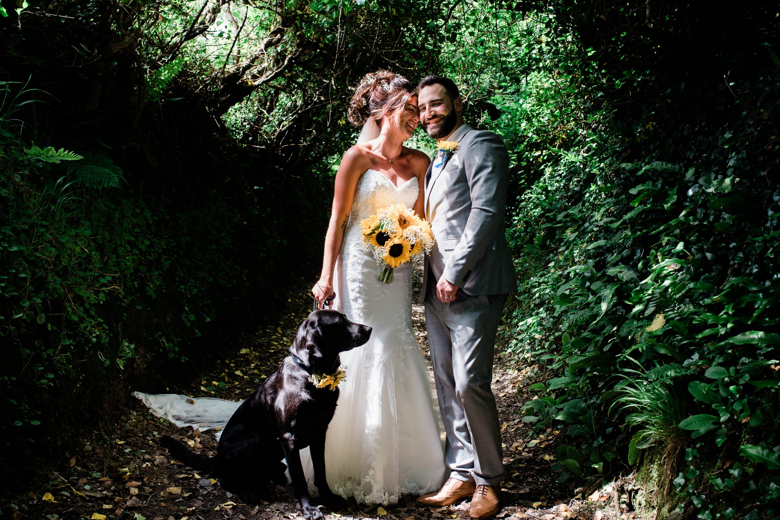 A bride and groom on their wedding day standing in a sunlit forested area with green foliage. The bride is wearing a white strapless wedding dress and holding a bouquet of sunflowers. The groom is wearing a light gray suit. They are smiling and leani
