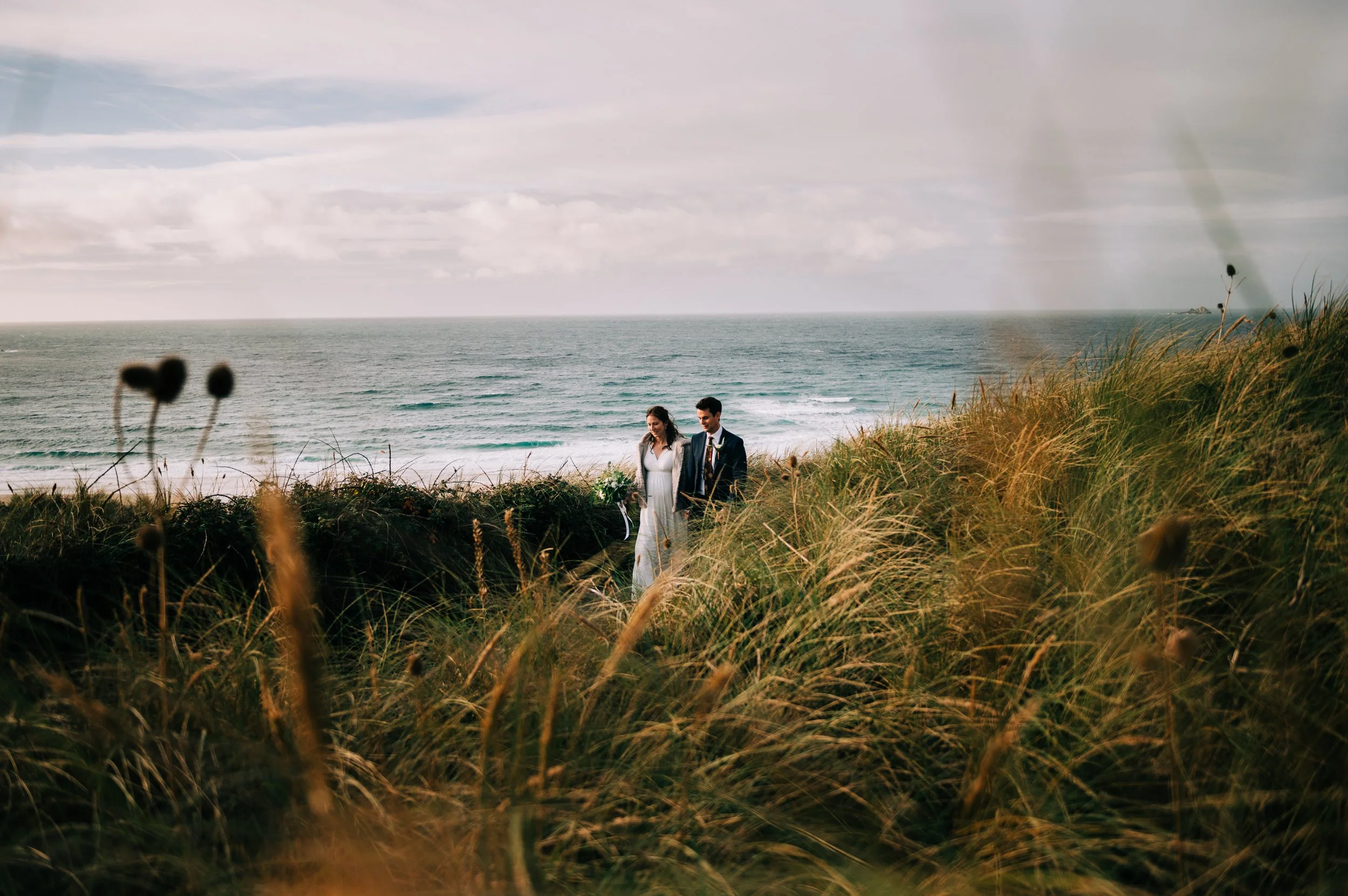 A couple dressed in wedding attire standing on a grassy dune by the ocean, with the sea and a cloudy sky in the background.