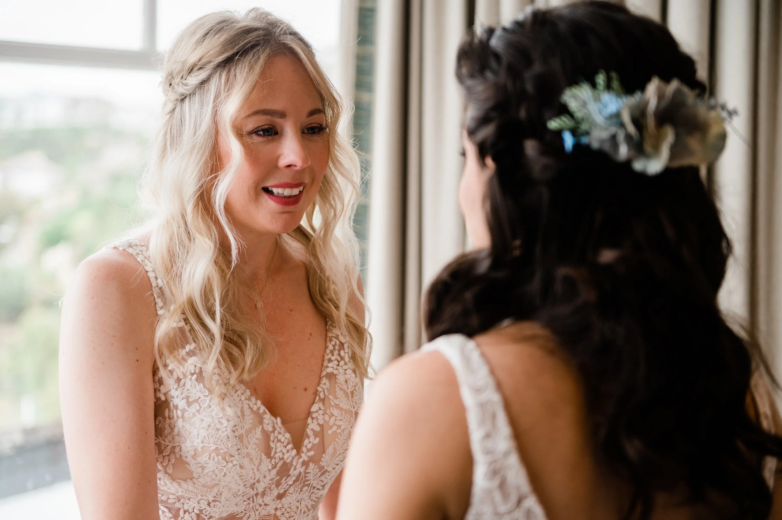 Two women, one with light blonde hair and one with dark brown hair, wearing white dresses inside a room with curtains. The blonde woman is smiling and facing the other woman, who has a floral hair accessory.