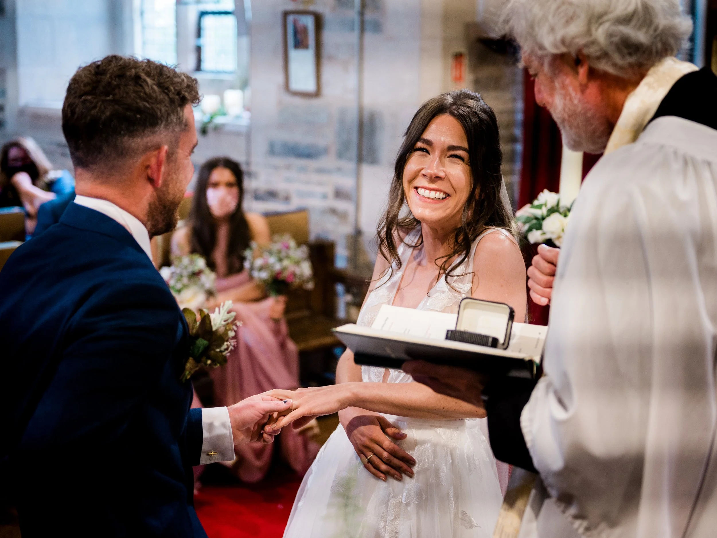 A bride and groom exchanging wedding rings during their ceremony, with the officiant holding a book, in a decorated indoor venue.