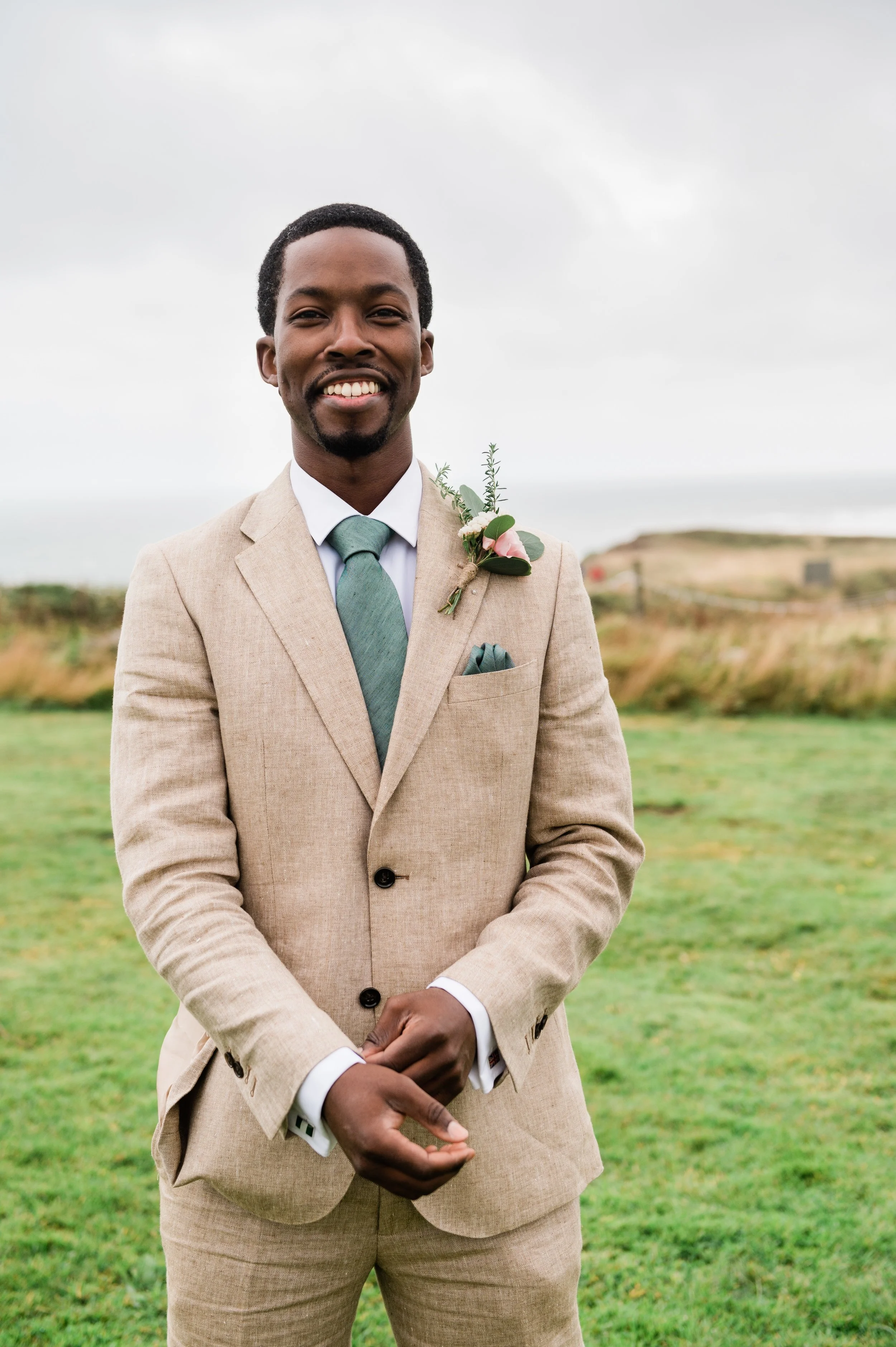 A smiling man in a beige suit and green tie standing outdoors on a grassy field, with a boutonniere pinned to his lapel.