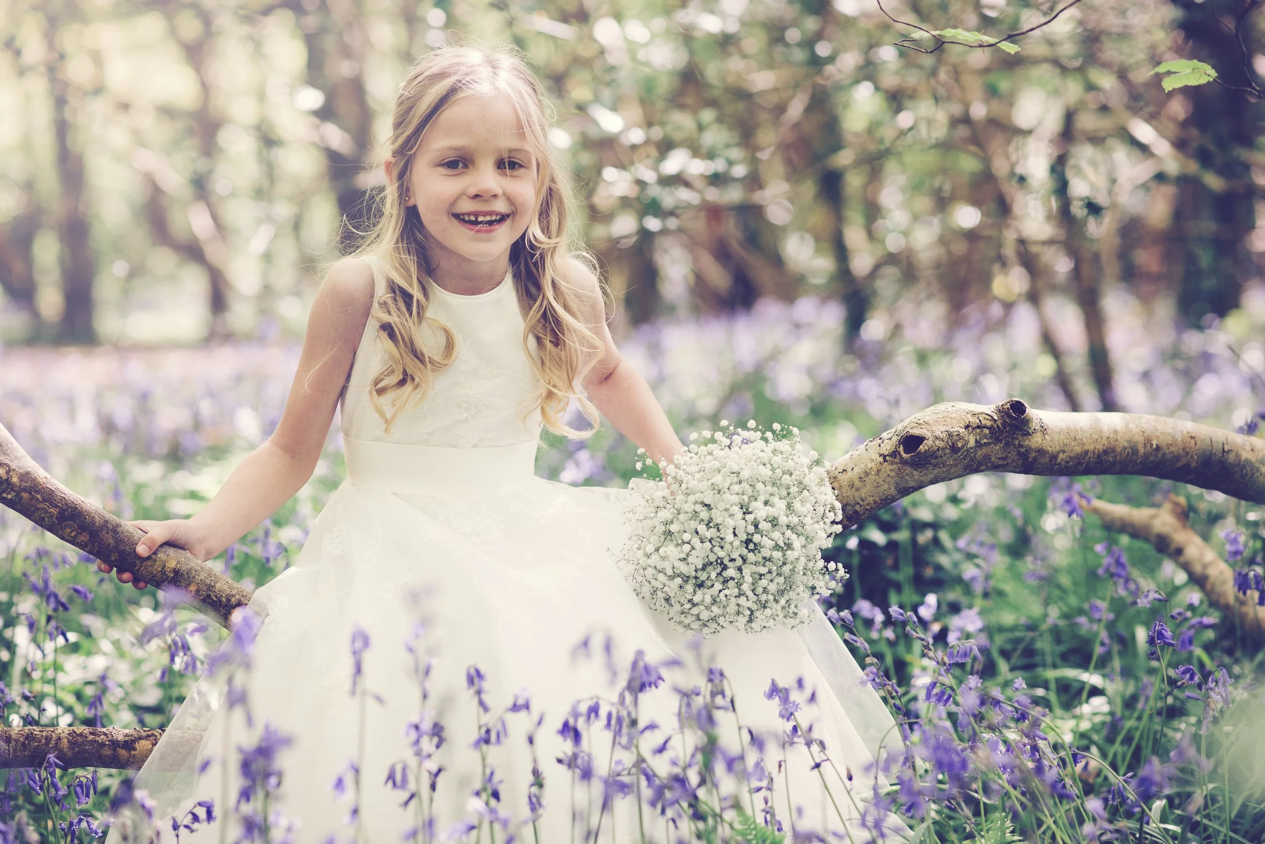 A young girl in a white dress sitting on a tree branch in a forested area with purple flowers, holding a bouquet of white flowers.