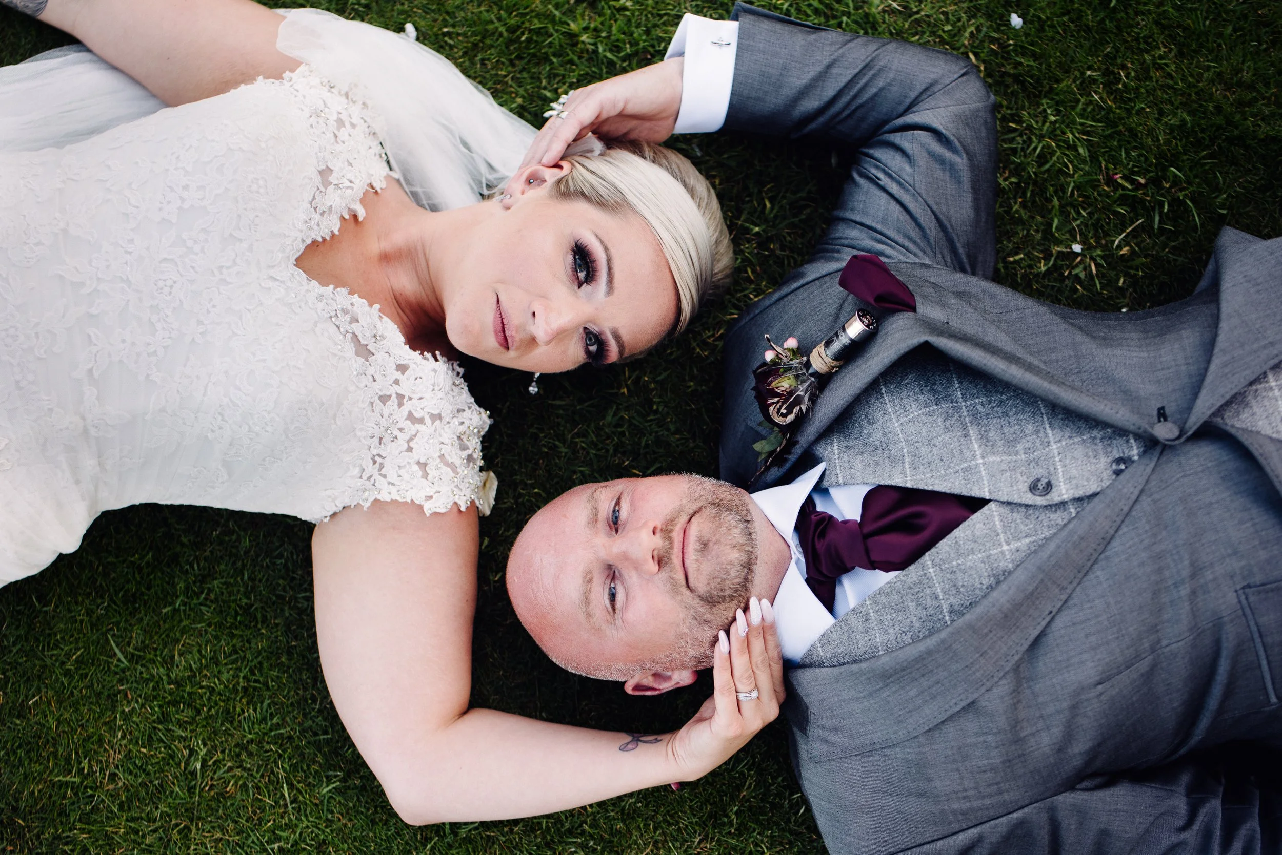 A bride and groom lying on grass, gazing at the camera, with the bride's hand resting on the groom's face.