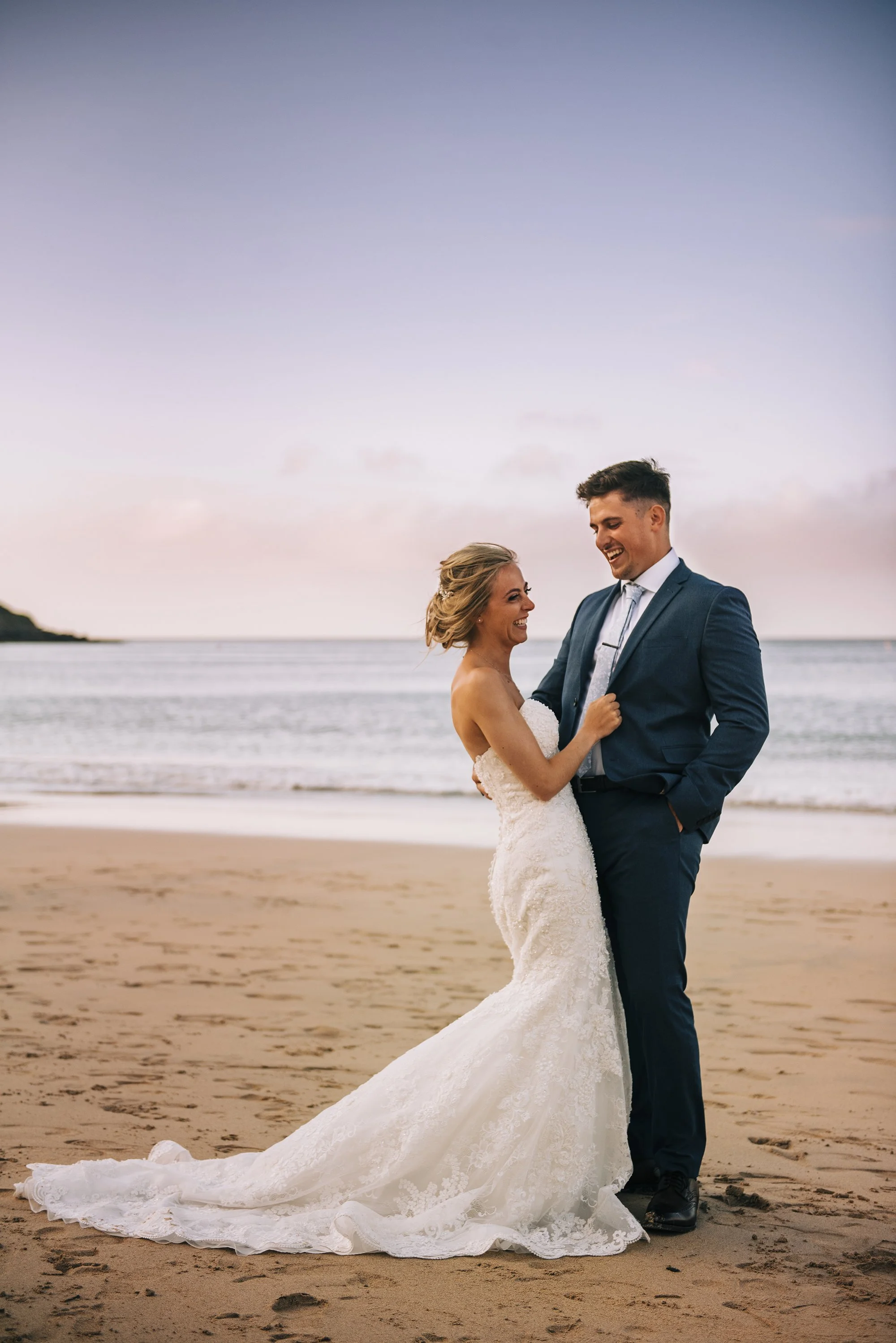 A newlywed couple in wedding attire on the beach at sunset, smiling and looking at each other.