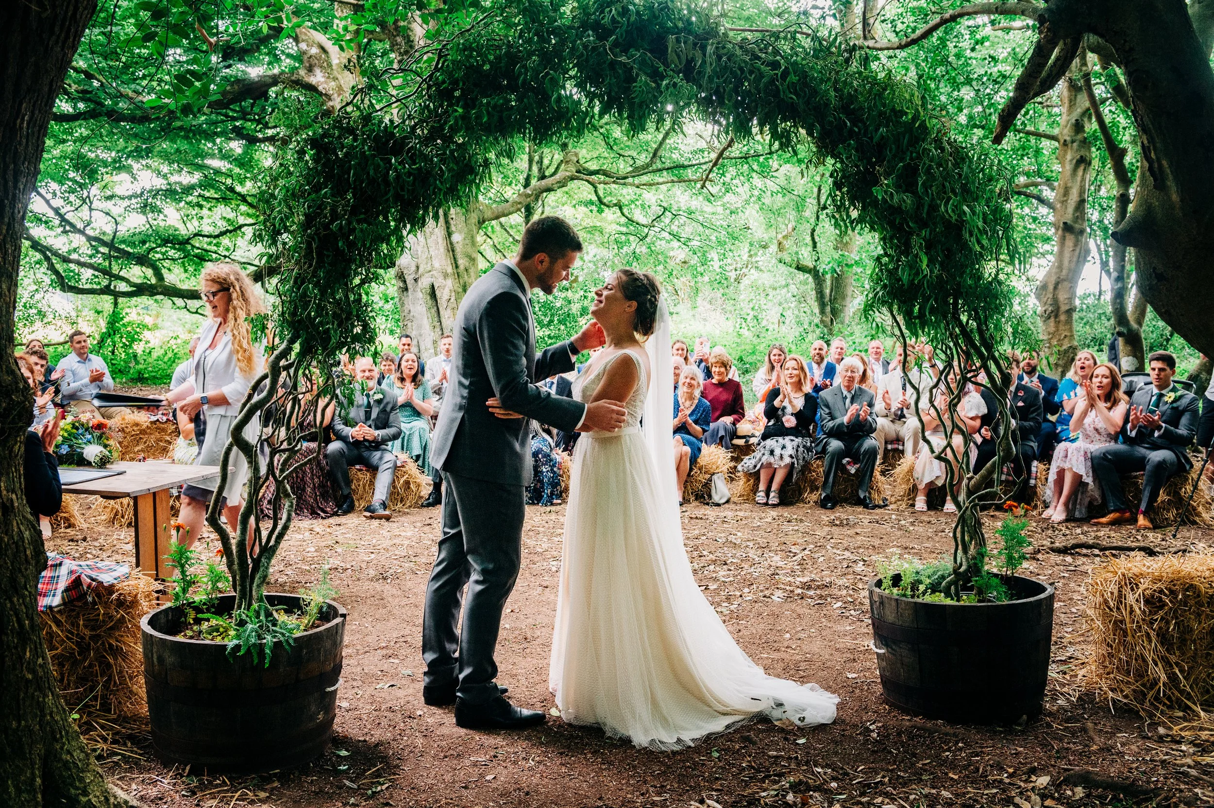 A couple getting married outdoors, standing under a leafy tree arch, surrounded by seated guests clapping and smiling.