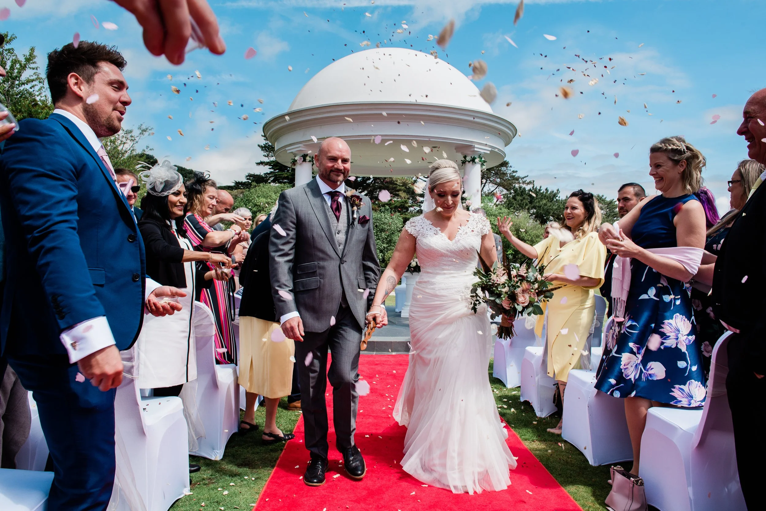 A bride and groom walking down the red carpet at their outdoor wedding ceremony with guests throwing confetti and smiling. The ceremony is set against a gazebo and the sky is blue with some clouds.