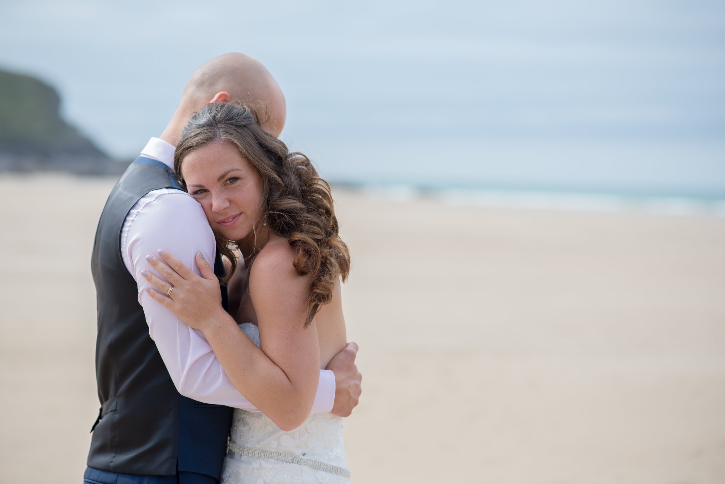 A newlywed couple embracing on a beach, with the bride looking at the camera and the groom with his head resting on her shoulder.