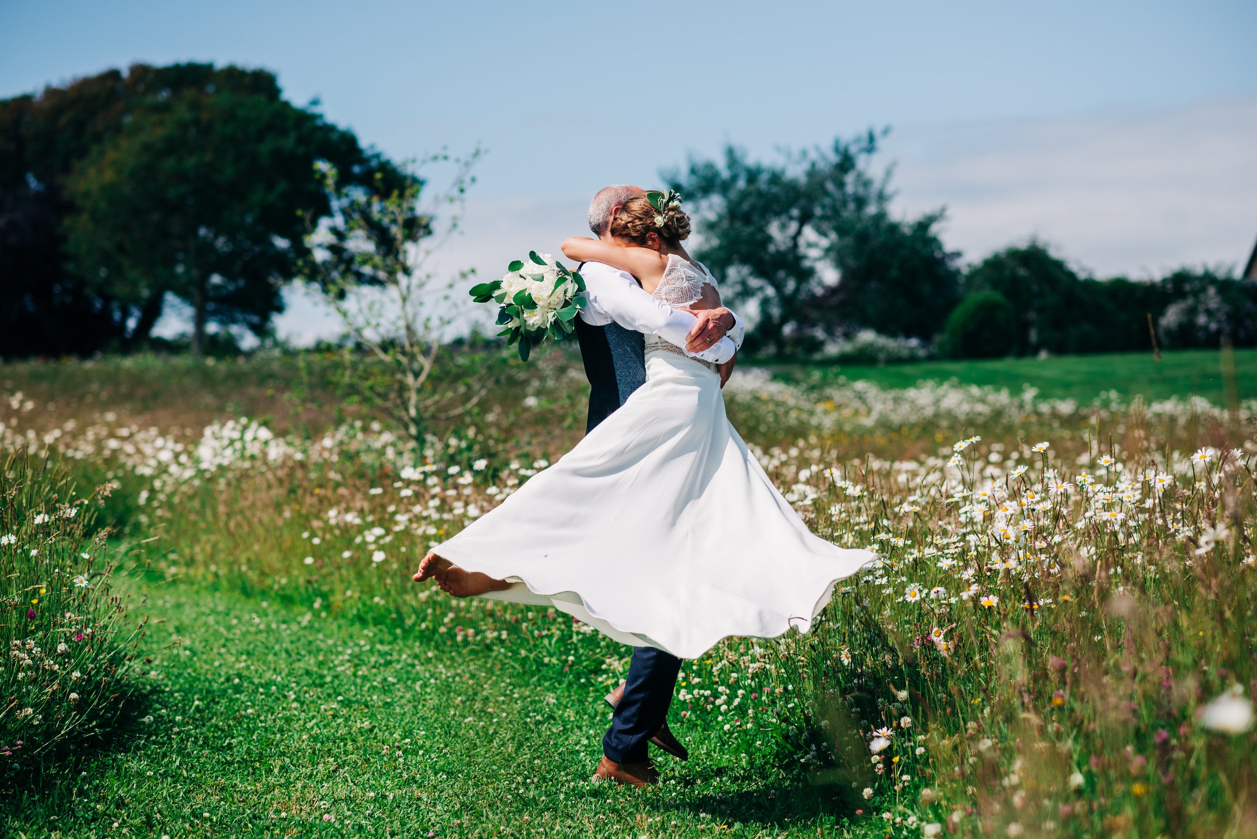 A bride and groom embracing in a field of flowers, with the groom lifting the bride off the ground in a joyful hug. The bride is holding a bouquet of white flowers.