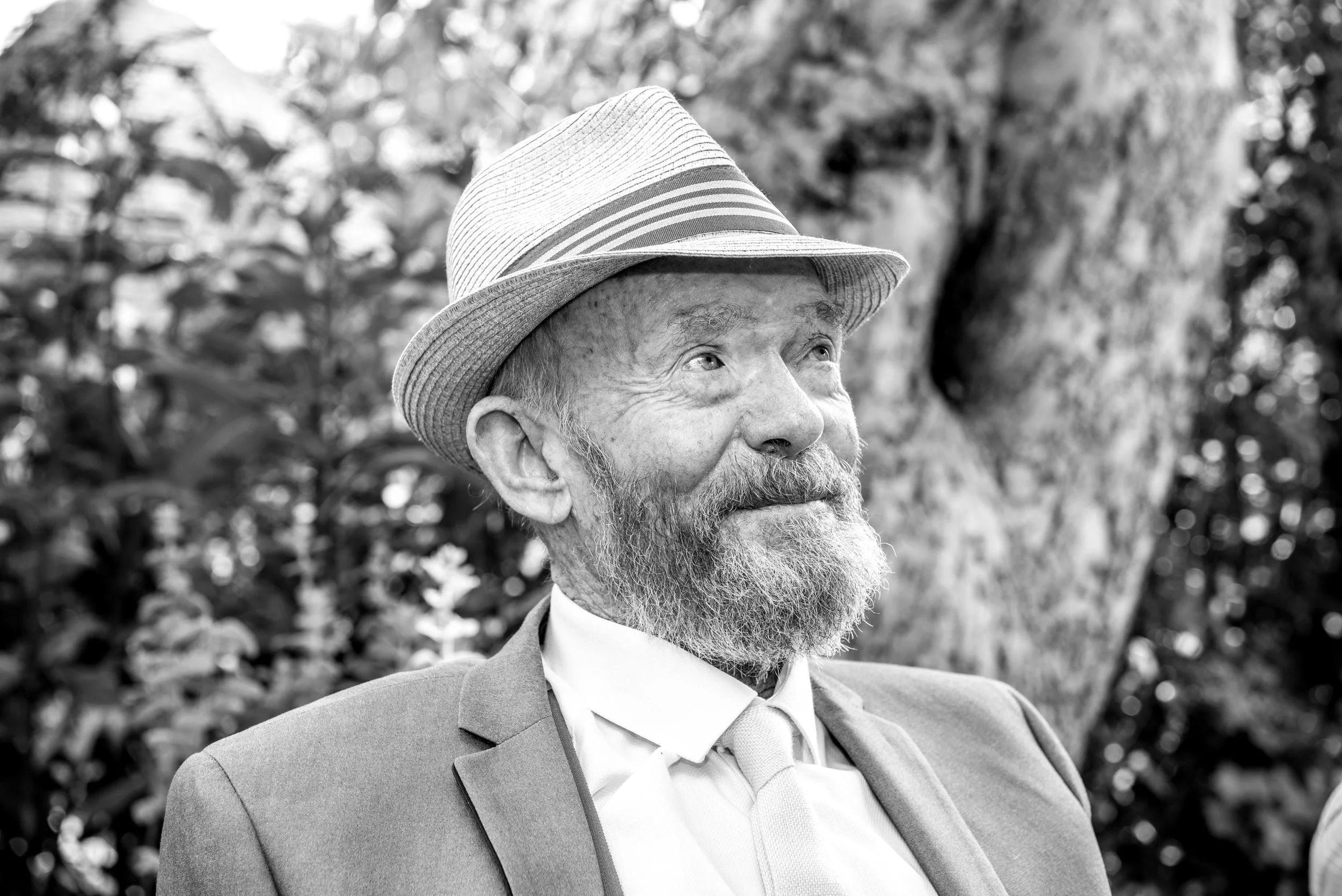 A black-and-white photo of an elderly man with a beard, wearing a fedora hat, suit, and tie, outdoors with blurred trees and foliage in the background.