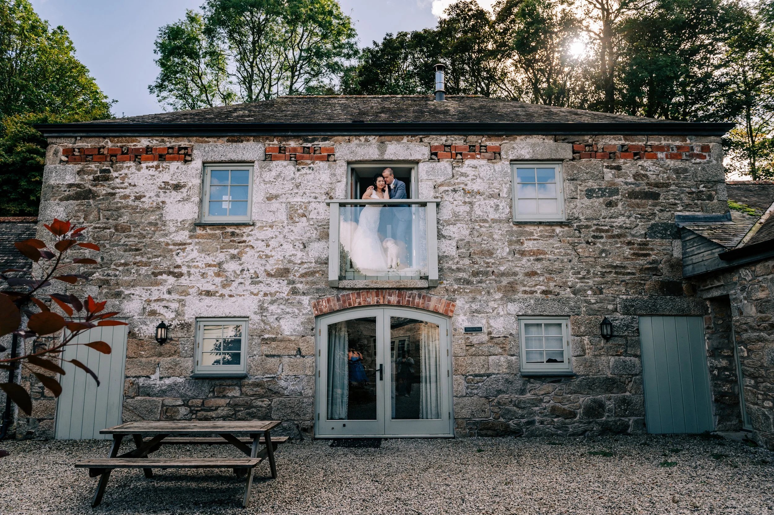 A stone house with a couple on the second-floor balcony, the bride in a wedding dress and veil, and the groom in a suit, inside the house. The house has two small windows on either side of the balcony, a large glass door below, and a picnic table out