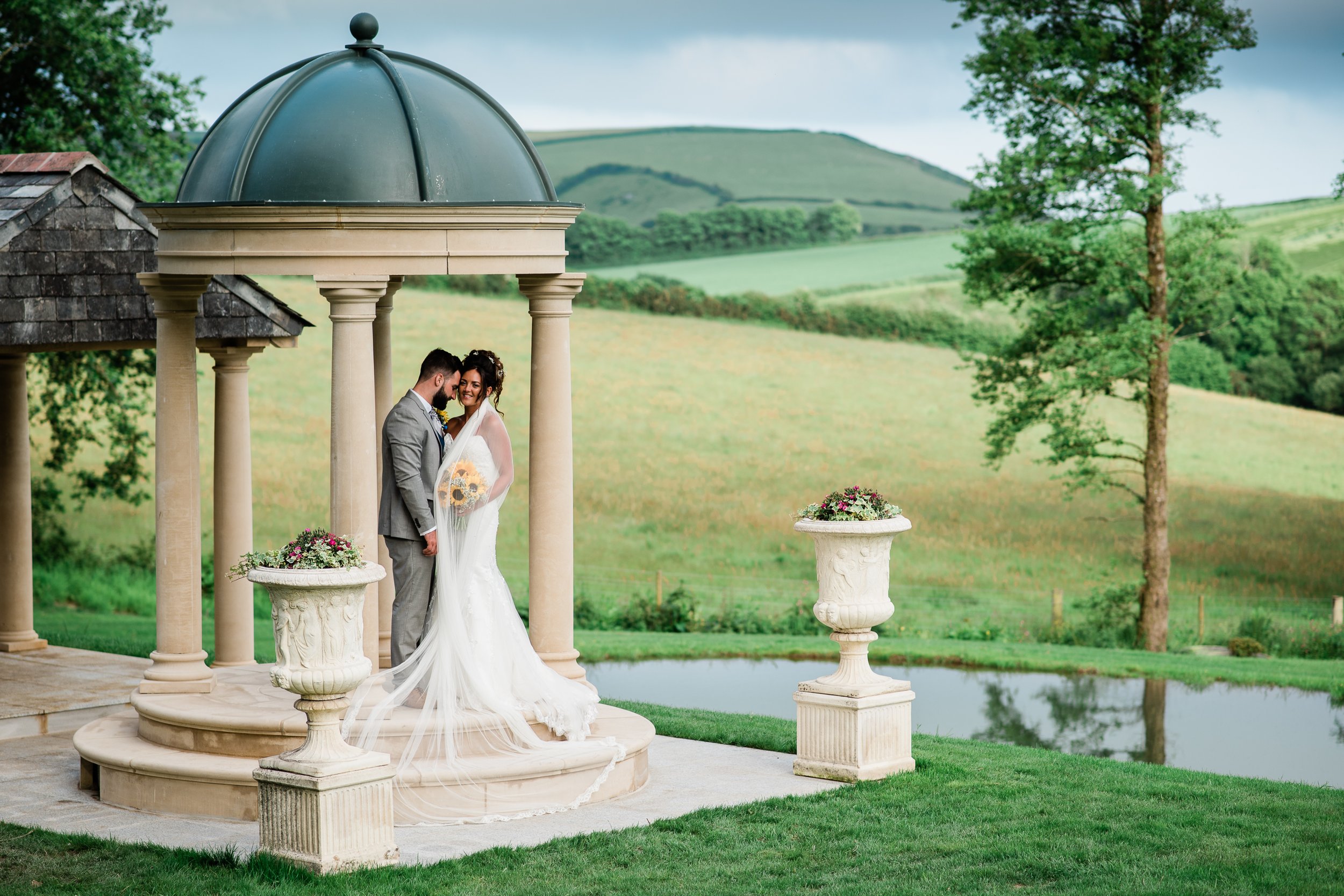 A bride and groom standing together on a marble platform with a domed gazebo, greenery, and rolling hills in the background on a cloudy day.