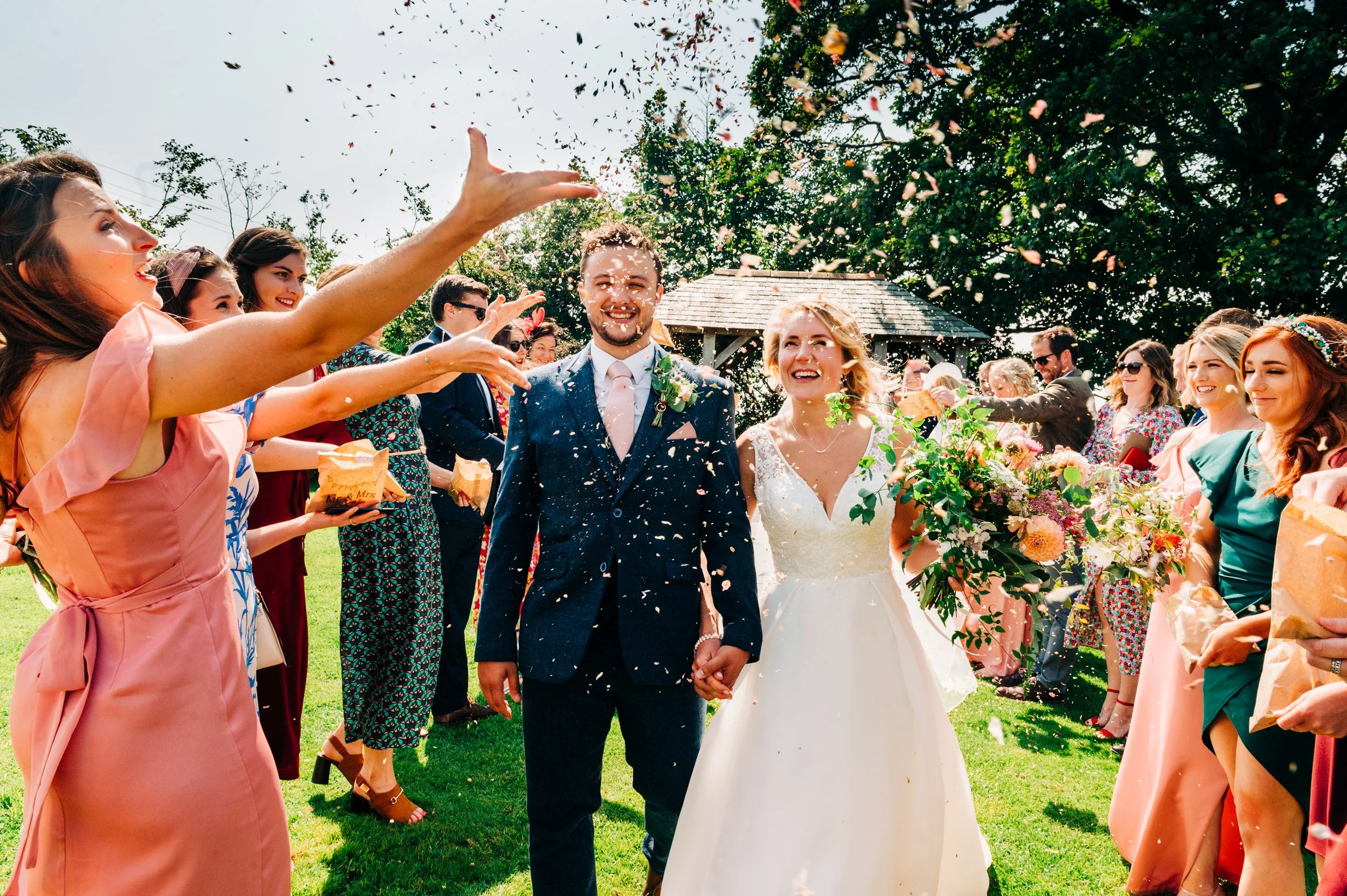 A newlywed couple walking hand in hand outdoors, surrounded by friends and family throwing flower petals and celebrating their wedding amidst a lush green garden under a bright, sunny sky.