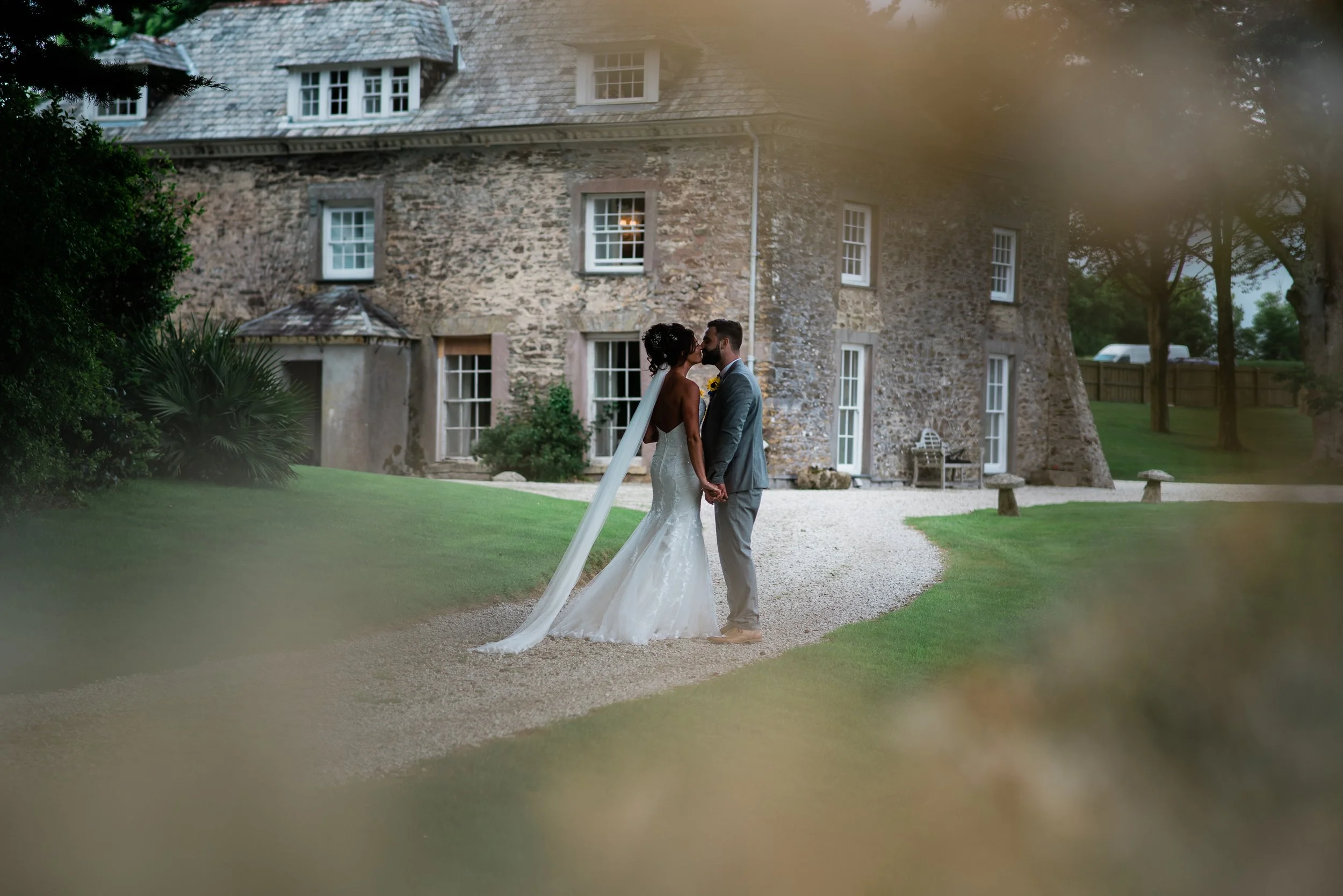A bride and groom standing close together outdoors in front of a stone building, holding hands and about to kiss, with greenery and trees around.