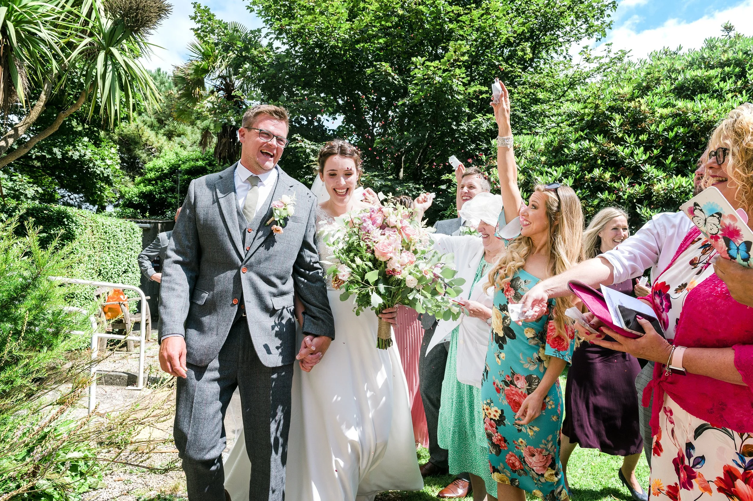 A wedding celebration outdoors with the bride and groom holding hands and smiling, surrounded by family and friends, some holding glasses, in a lush garden setting under a bright sky.