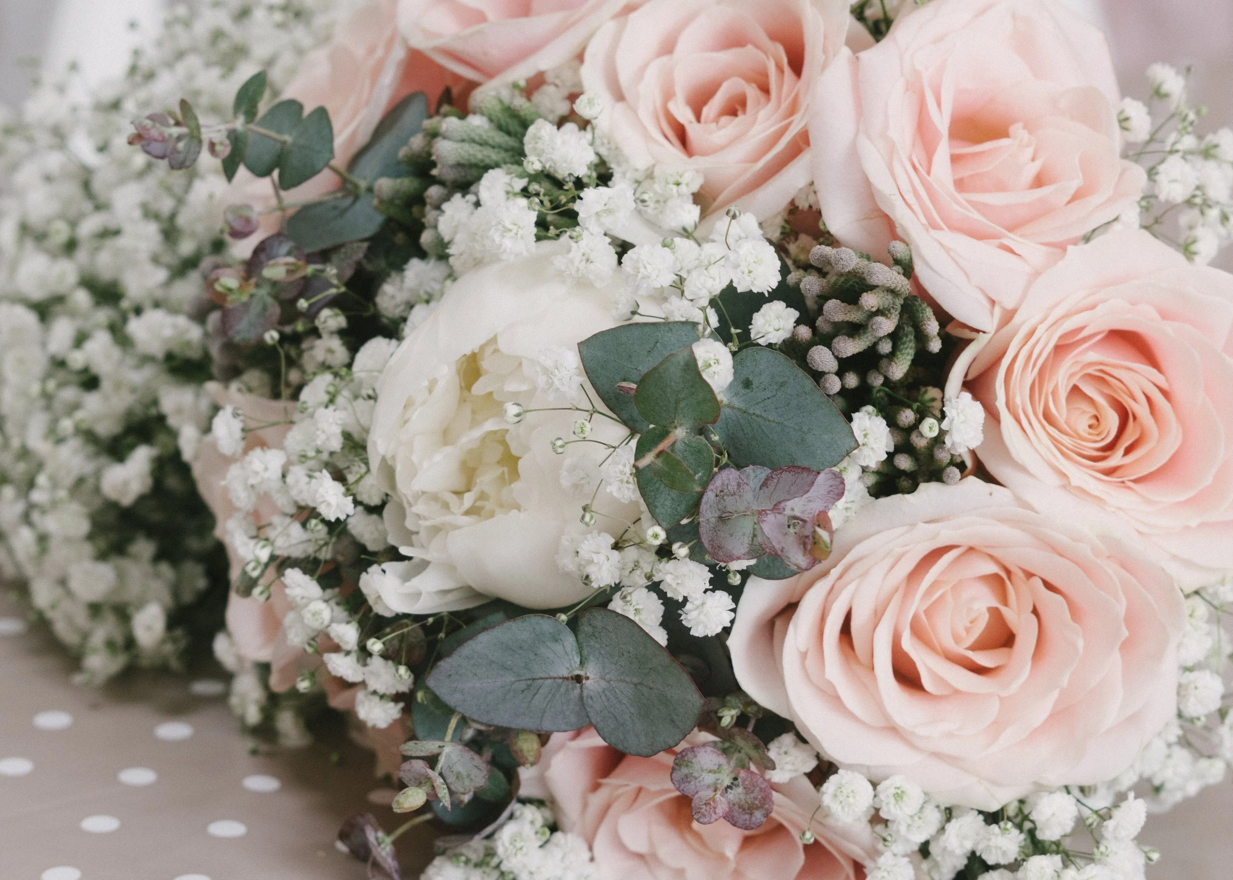 Close-up of a bouquet with light pink roses, white peonies, baby's breath, eucalyptus leaves, and other small flowers.