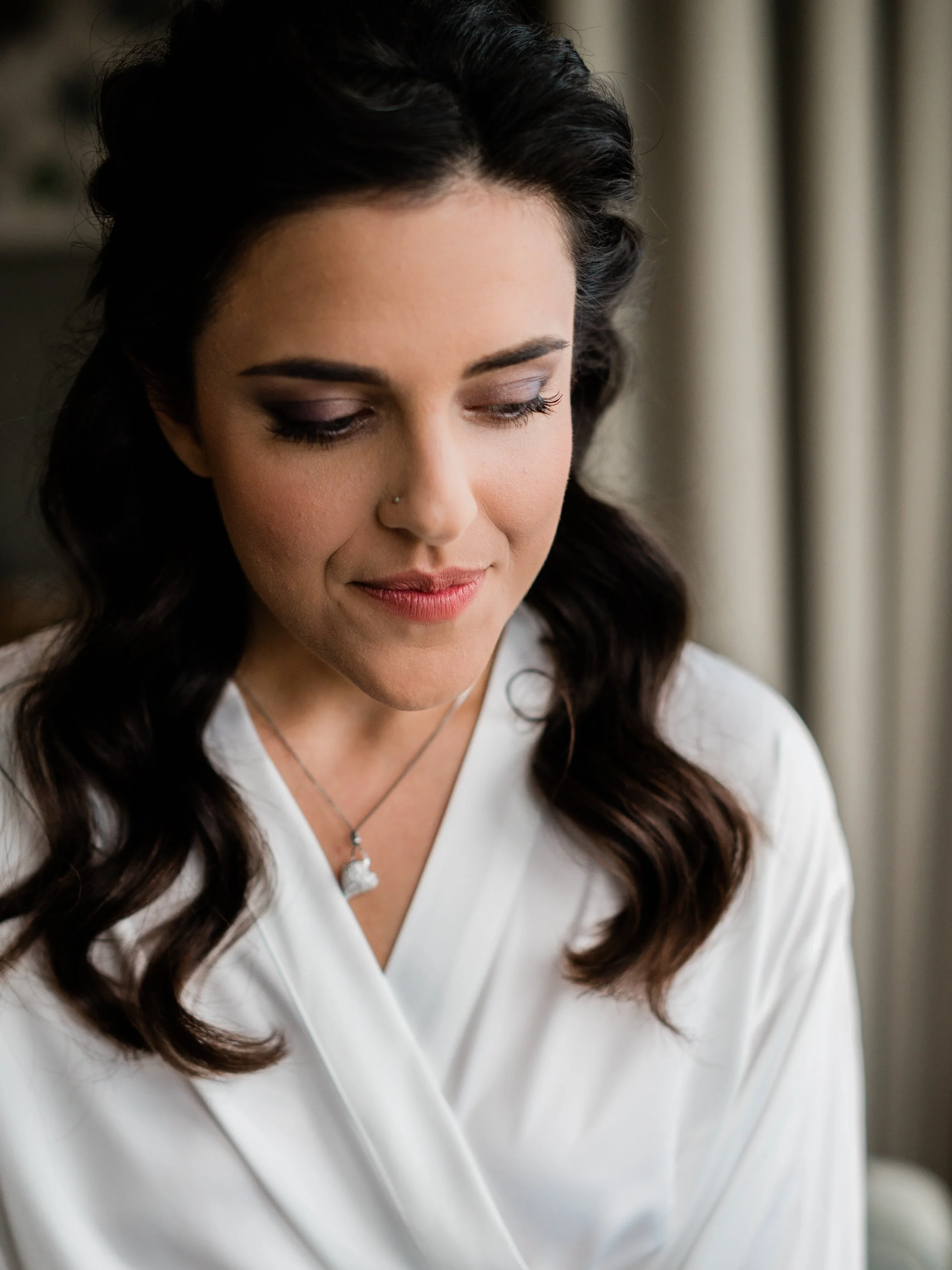 A woman with dark hair styled in loose waves, wearing a white satin robe, looking down with a gentle smile, wearing a necklace with a heart-shaped pendant, in a softly lit room with beige curtains.