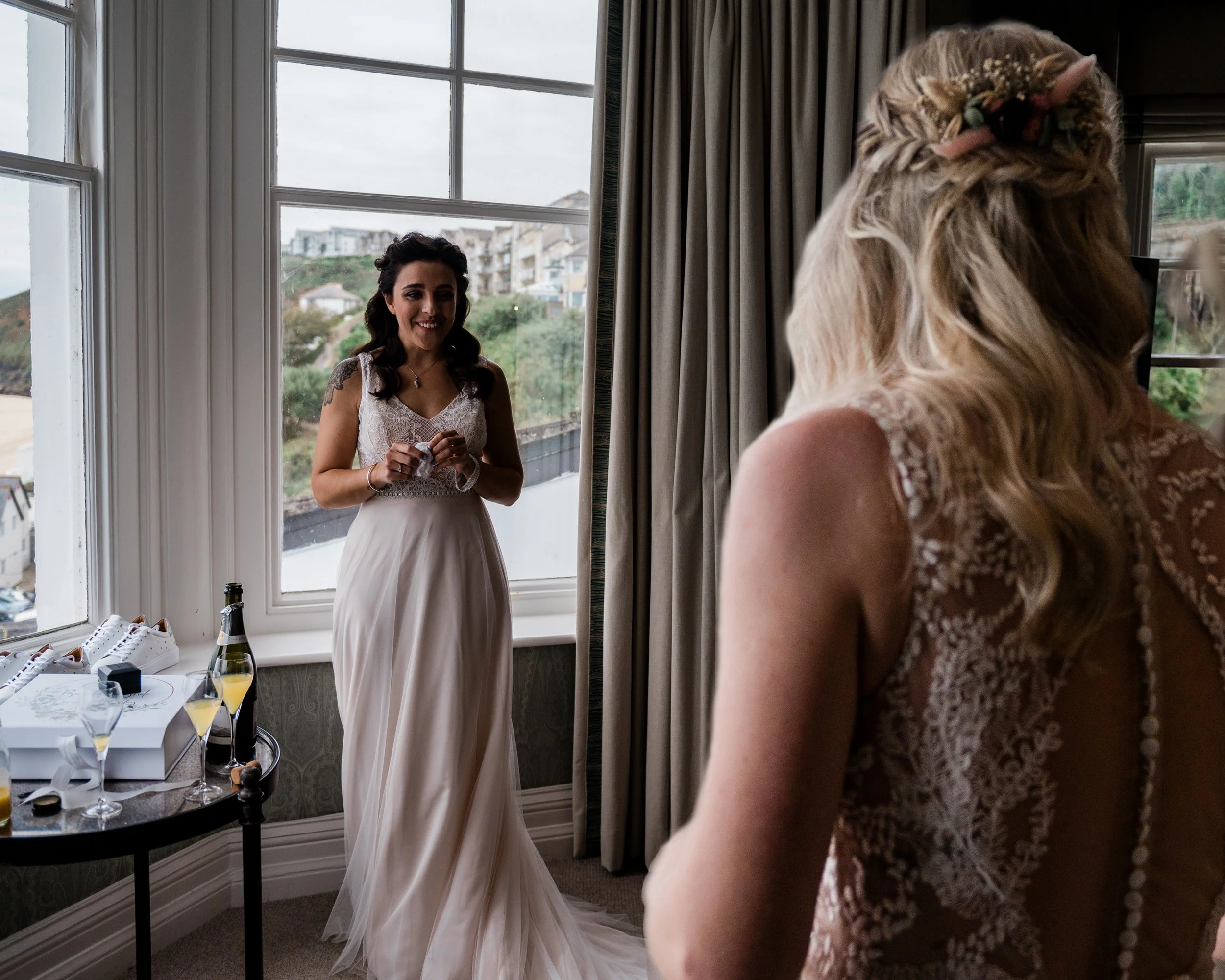 A bride in a white wedding gown standing in front of a window, smiling, with a woman in a lace dress watching her.