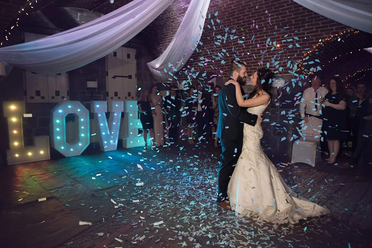 Couple dancing at a wedding reception surrounded by confetti, with large illuminated letters spelling 'LOVE' in the background.