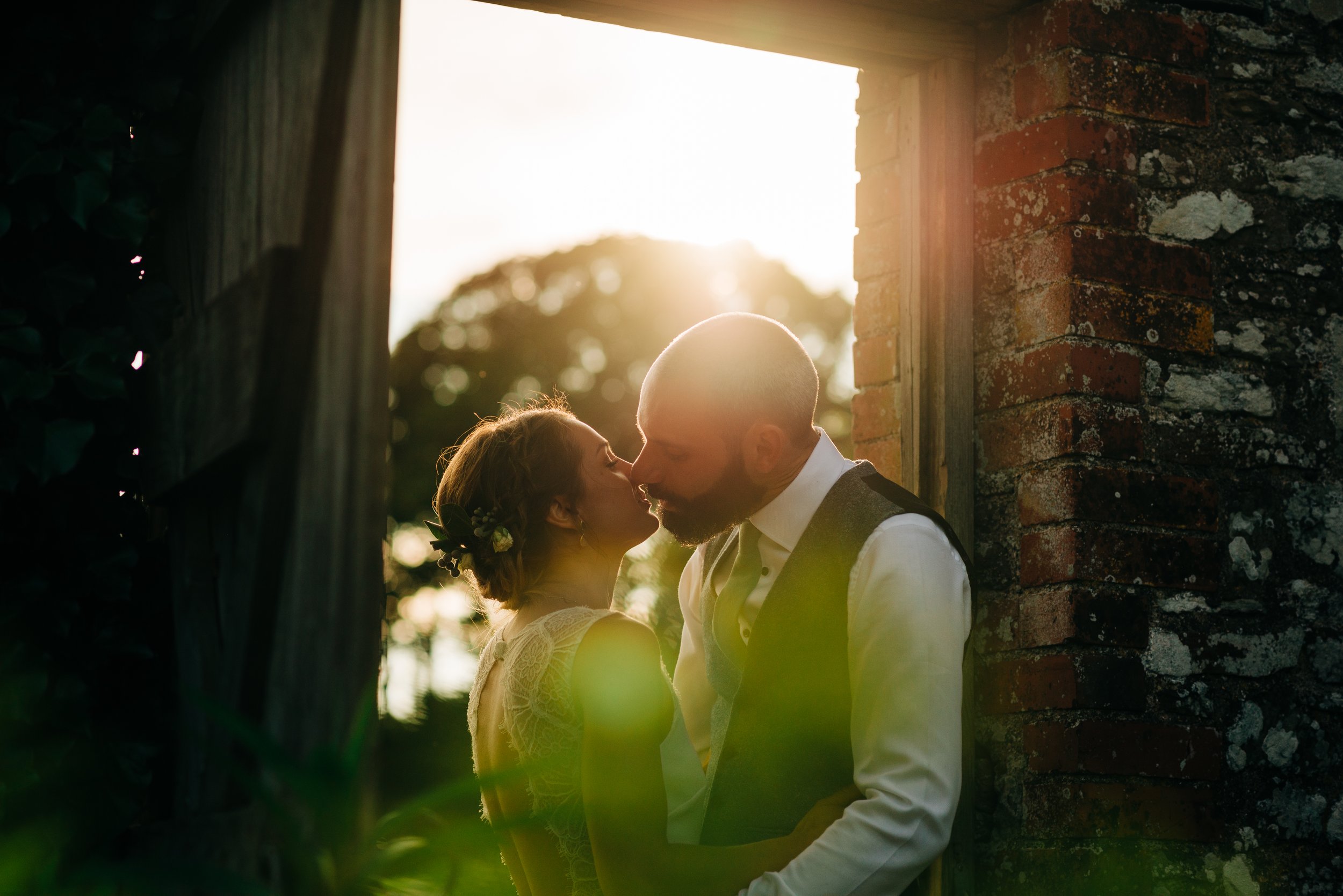 A couple is kissing in front of a rustic brick and wood structure with sunlight shining behind them, outdoors during sunset.